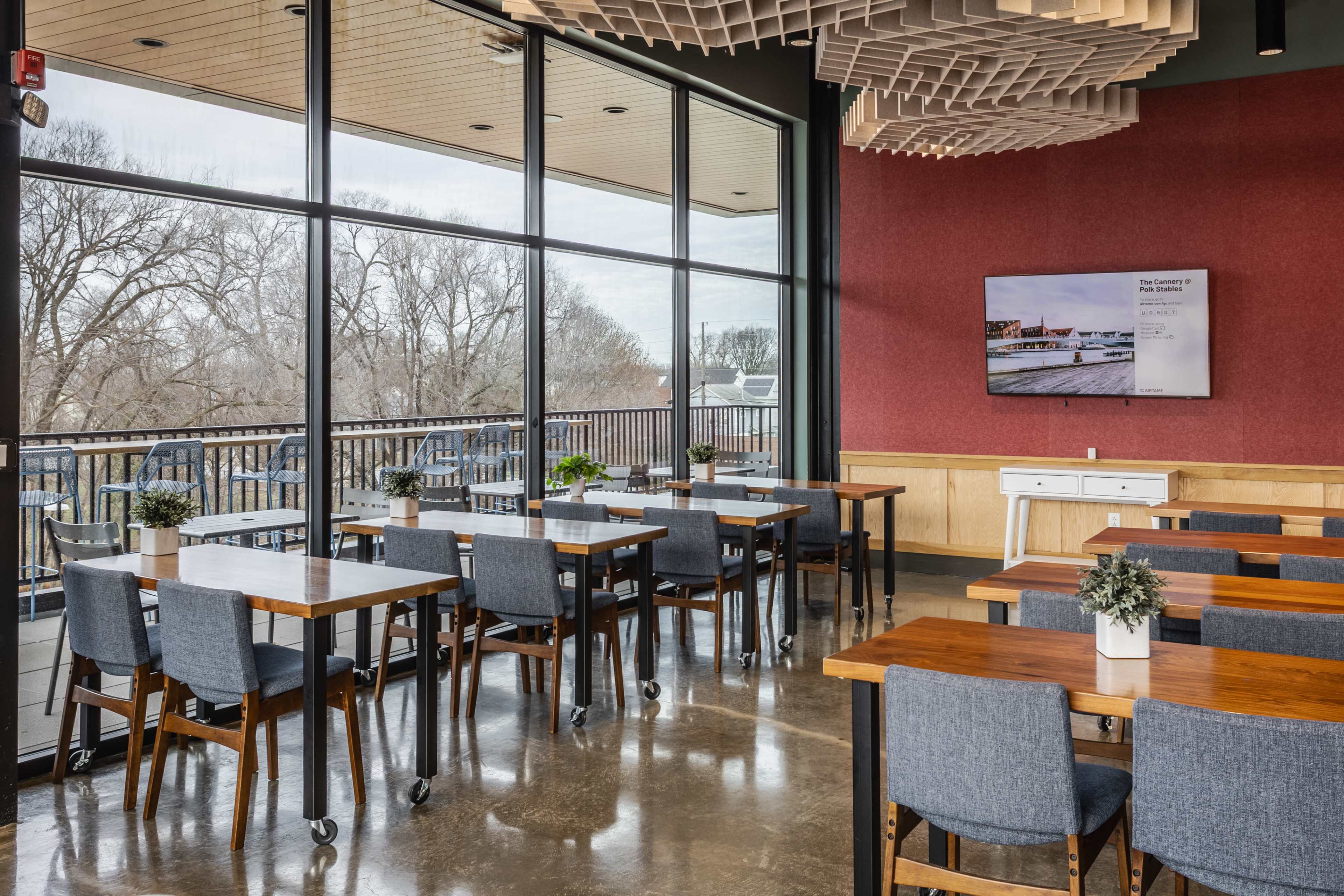 A modern meeting room features multiple wooden tables with chairs, large windows providing a view of trees outside, and a wall-mounted screen displaying information.