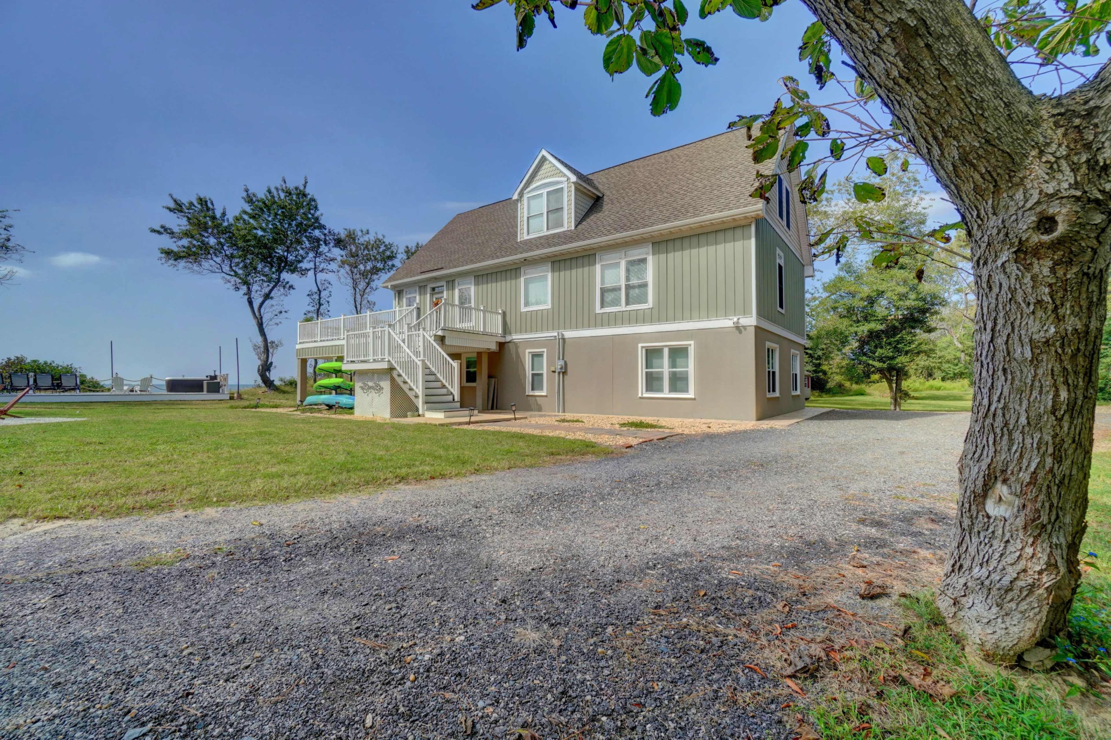 A two-story house with a gray exterior and a large driveway is situated in a grassy area with a few trees nearby.