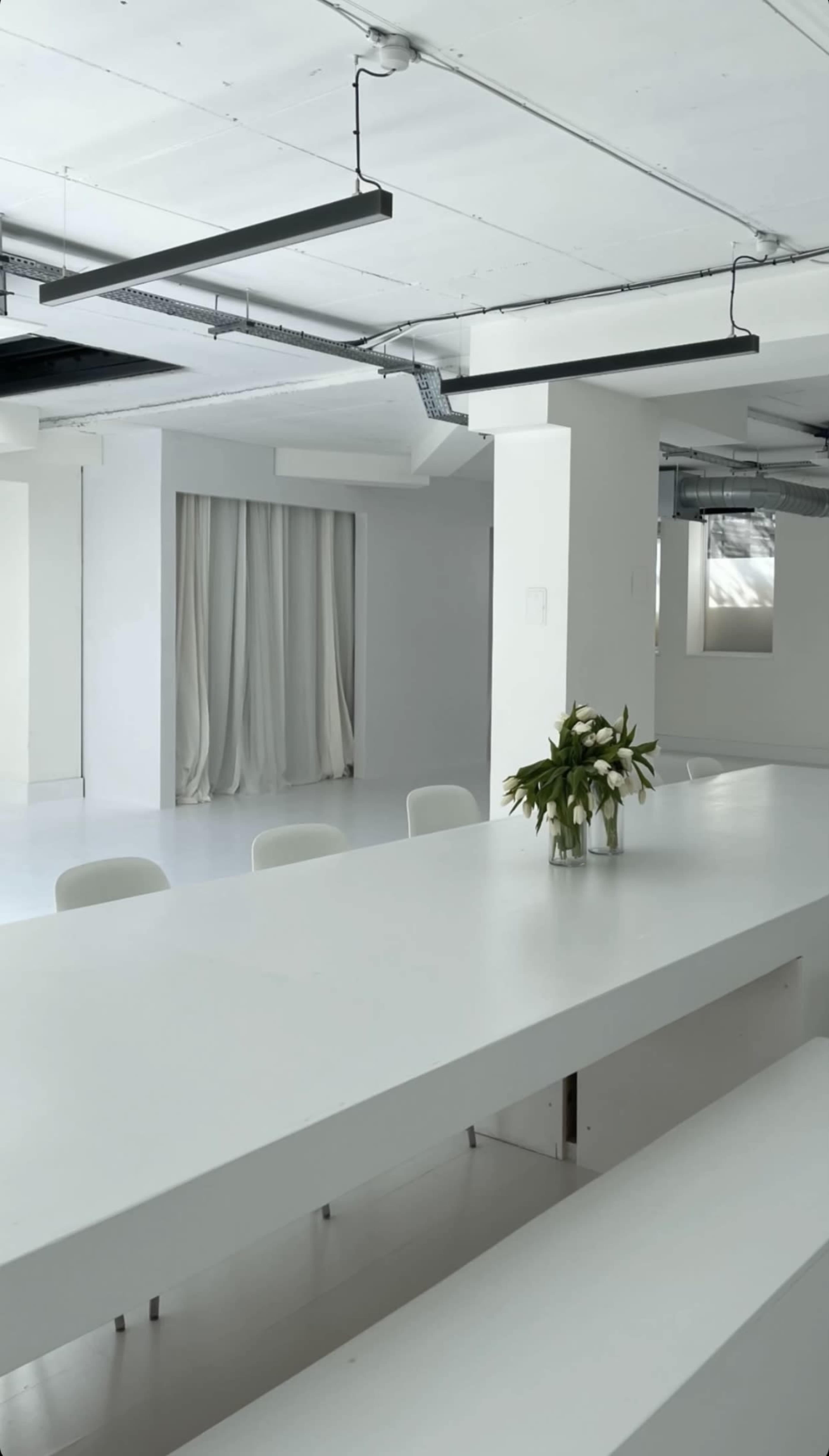 A minimalist white meeting room with a long table, white chairs, and a vase of flowers in the center.
