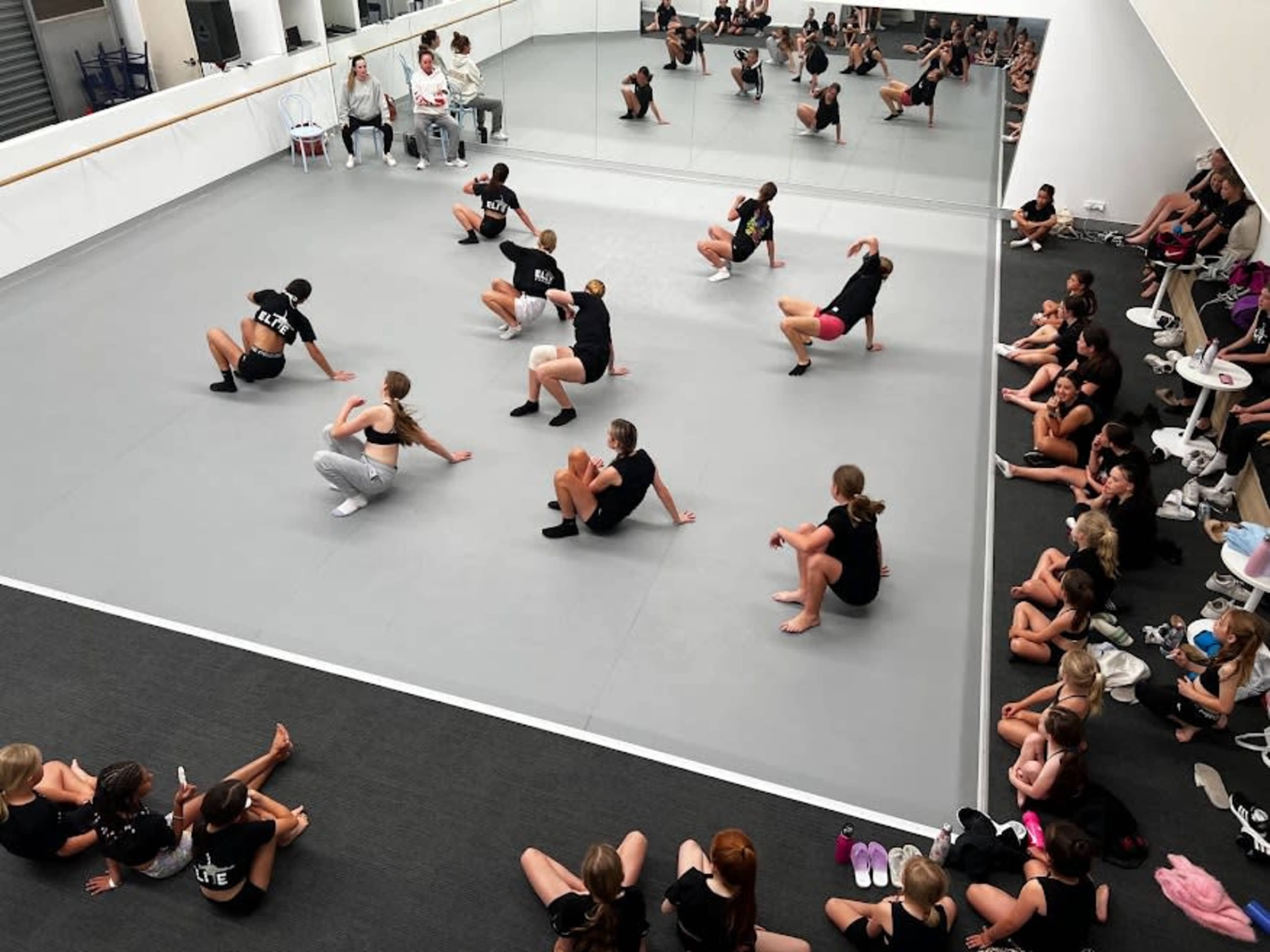 A group of dancers in black shirts practices in a mirrored studio while seated observers watch from the sidelines.