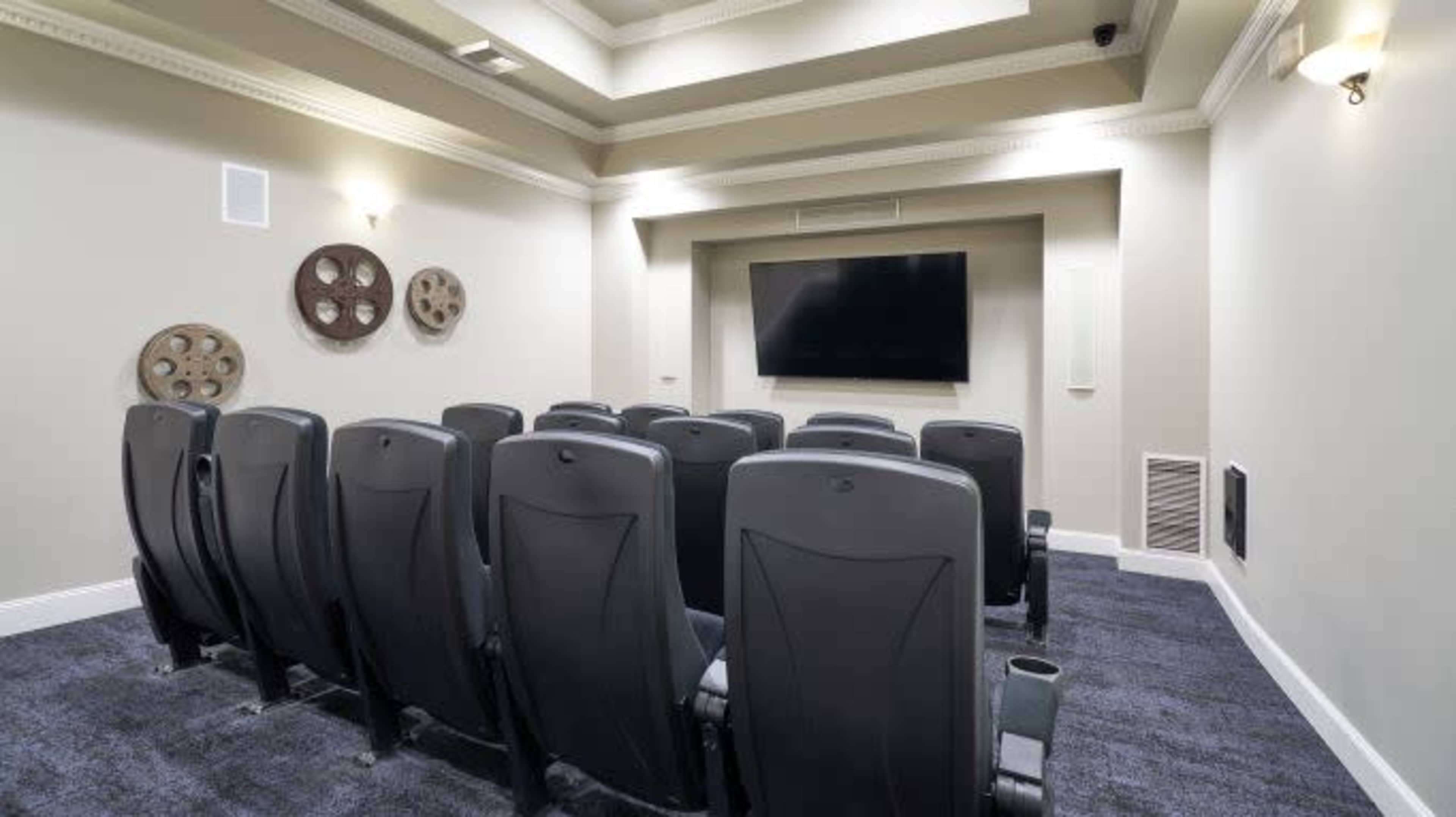 The image shows a small home theater with black seating arranged in rows facing a large flat-screen TV on a light-colored wall.
