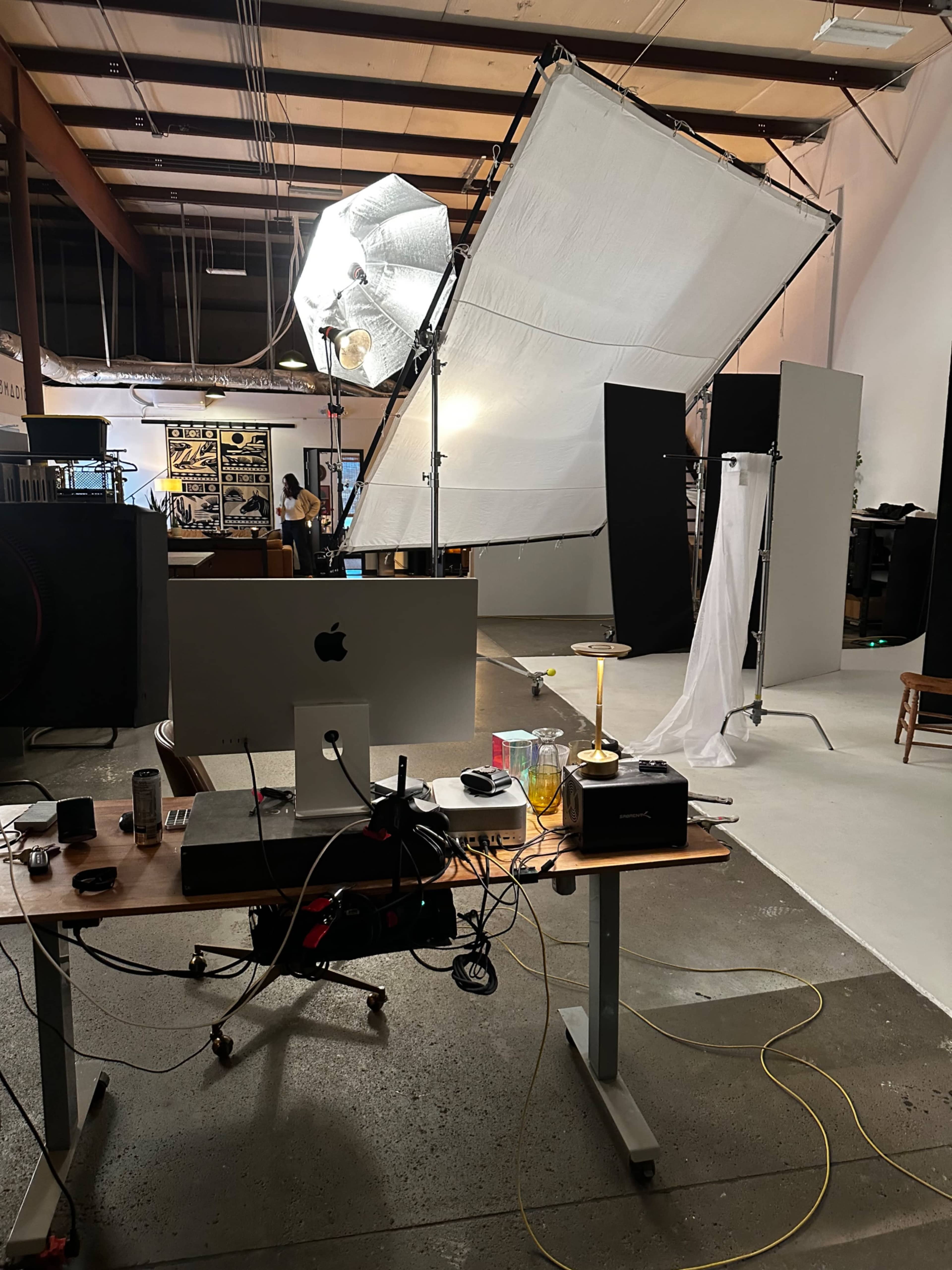 The image shows a photography studio with a desk cluttered with equipment in the foreground, a large softbox lighting setup, and a backdrop on the right.