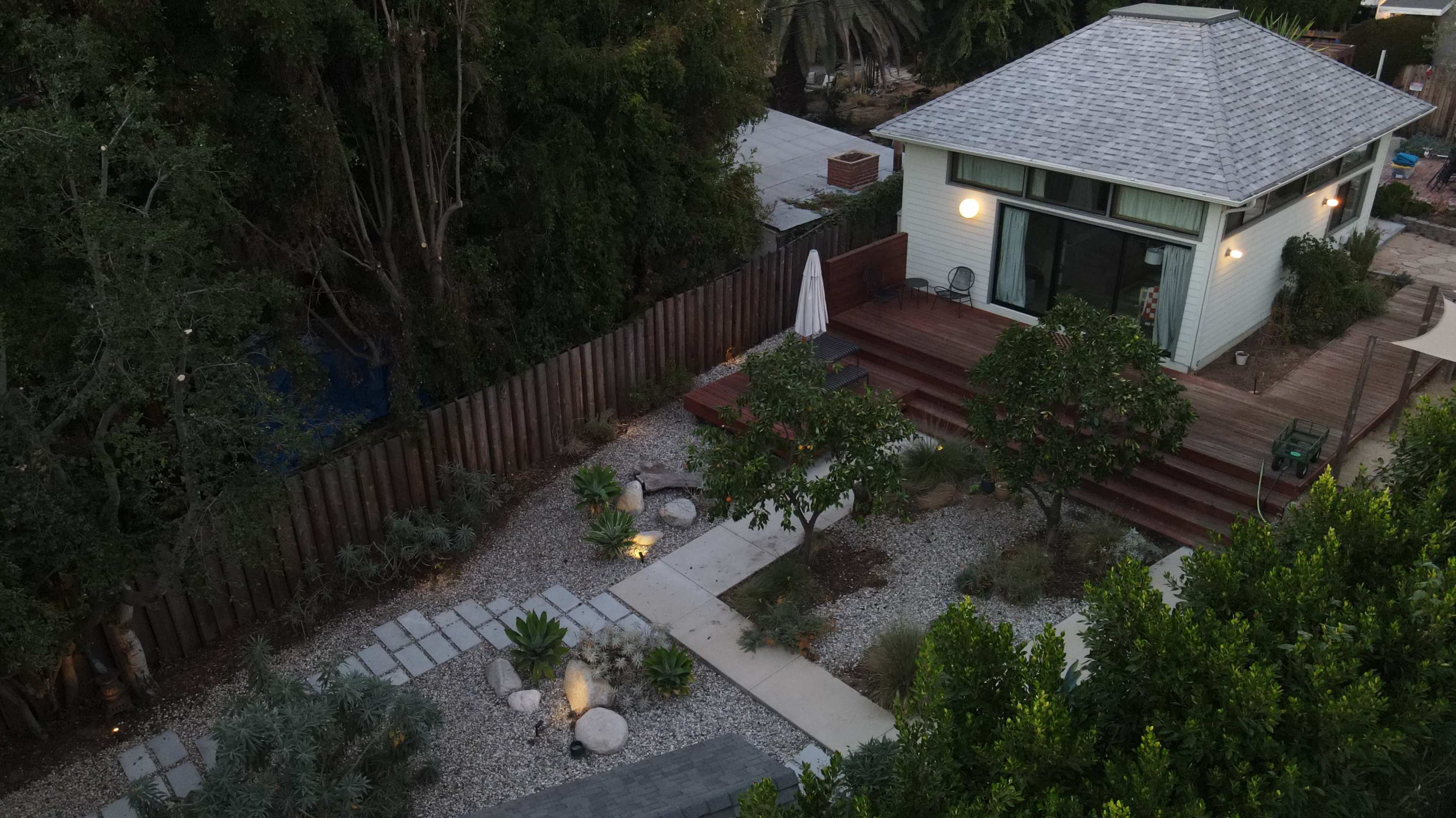 The aerial view shows a landscaped backyard with a wooden deck, pathways lined with gravel, and several trees, leading to a small house with a gabled roof.