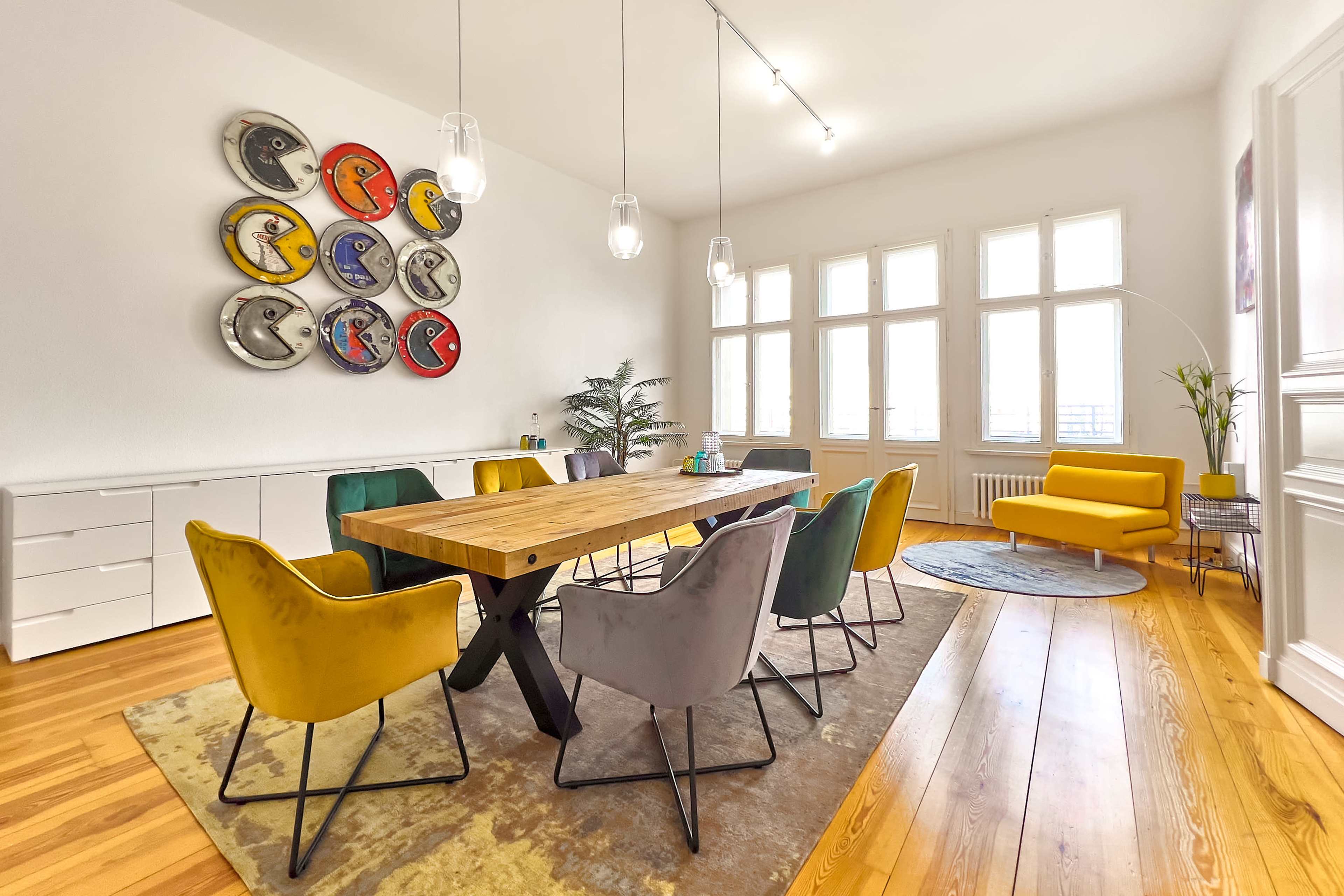 A modern dining area with a large wooden table surrounded by colorful chairs, a decorative wall featuring circular artworks, and natural light coming through several windows.