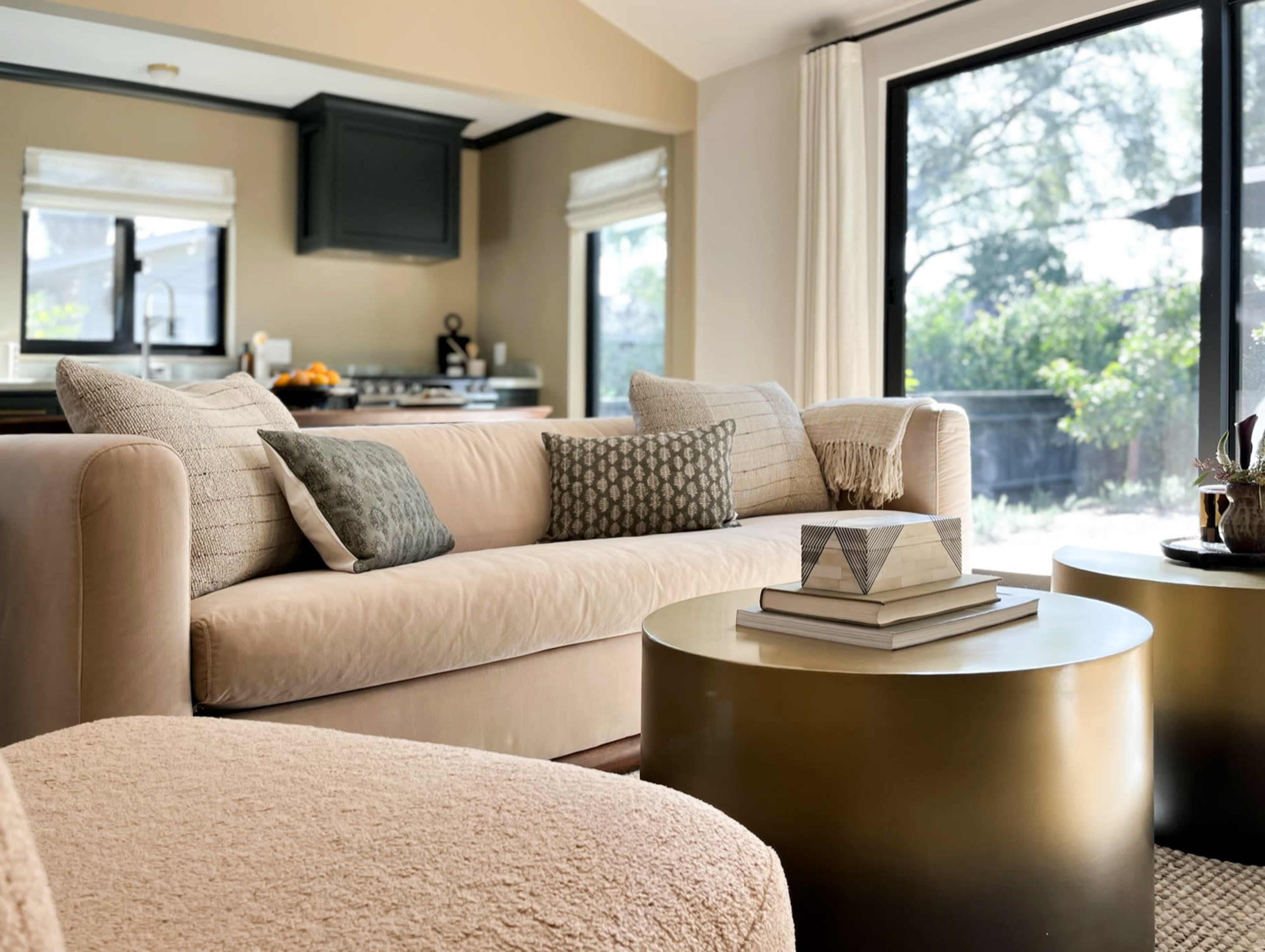 A living room with a beige sofa adorned with decorative pillows, gold side tables, and large windows allowing natural light to enter.