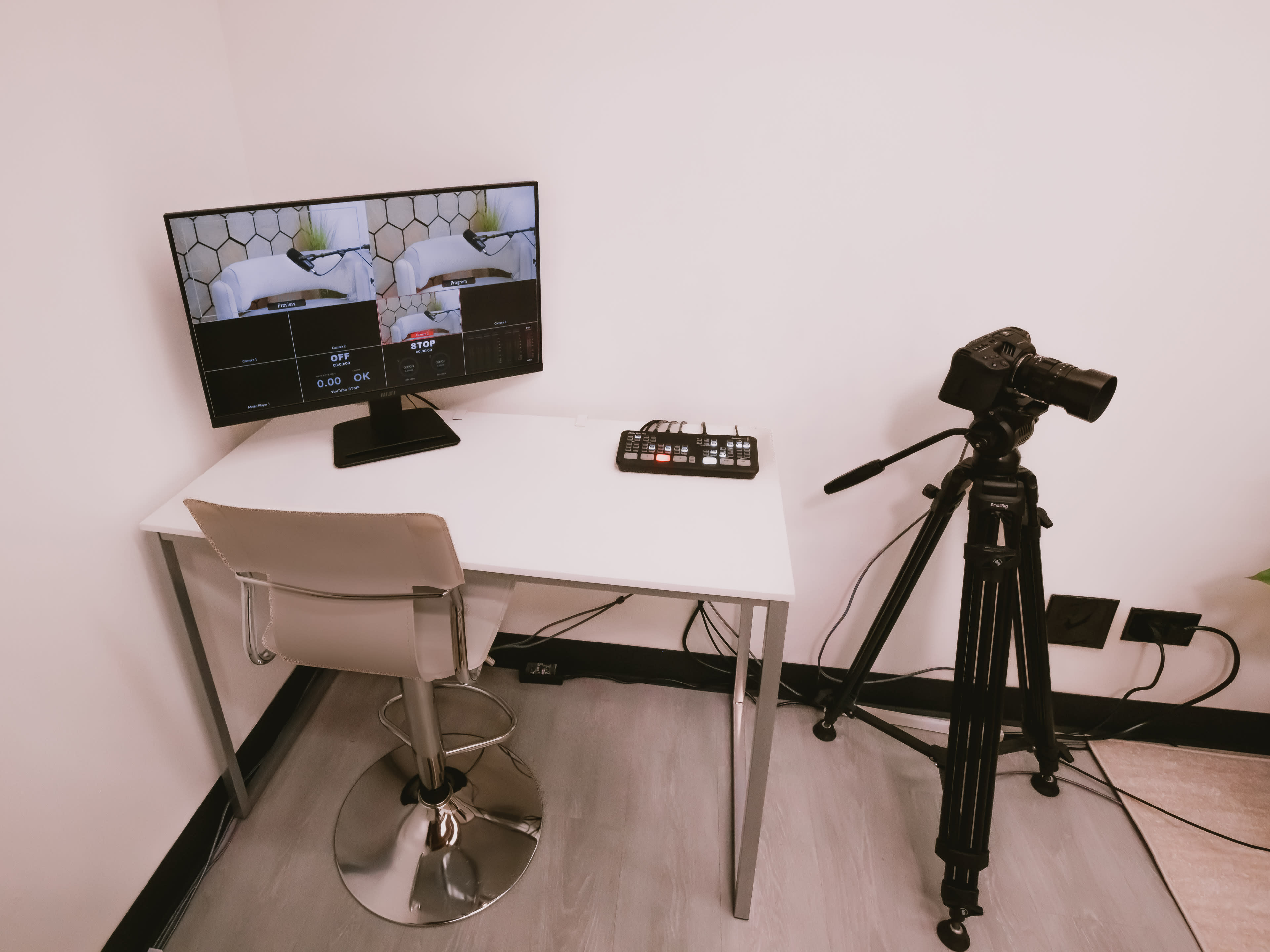 The image shows a minimalistic video production setup featuring a monitor displaying a camera feed, a microphone interface, and a camera mounted on a tripod beside a simple desk and chair.