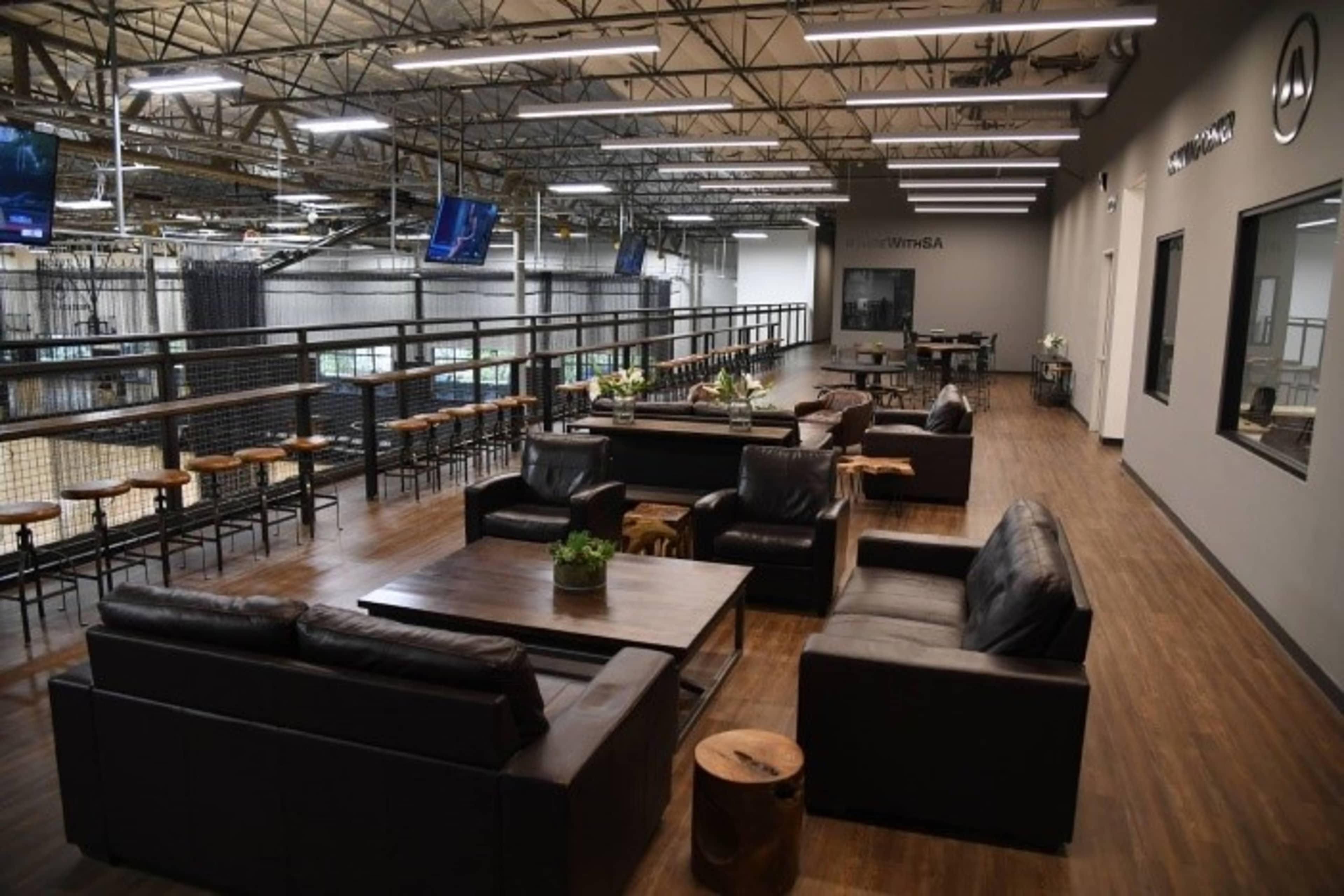 A spacious office area with leather couches, a wooden table, and a balcony overlooking a sports facility, complemented by large televisions mounted on the walls.