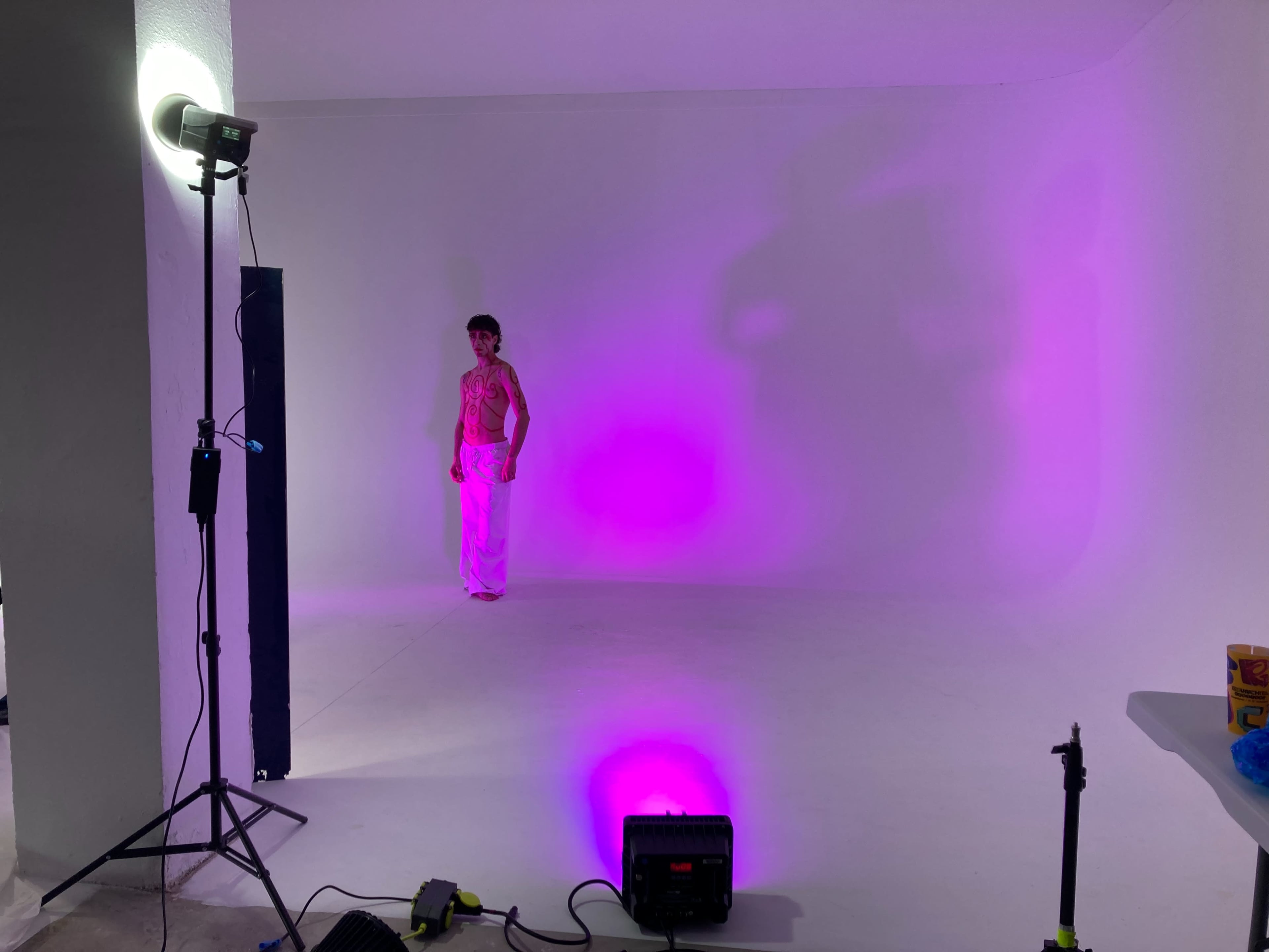 A person stands against a blank white wall illuminated by purple lighting, with photography equipment and cables arranged on the floor.
