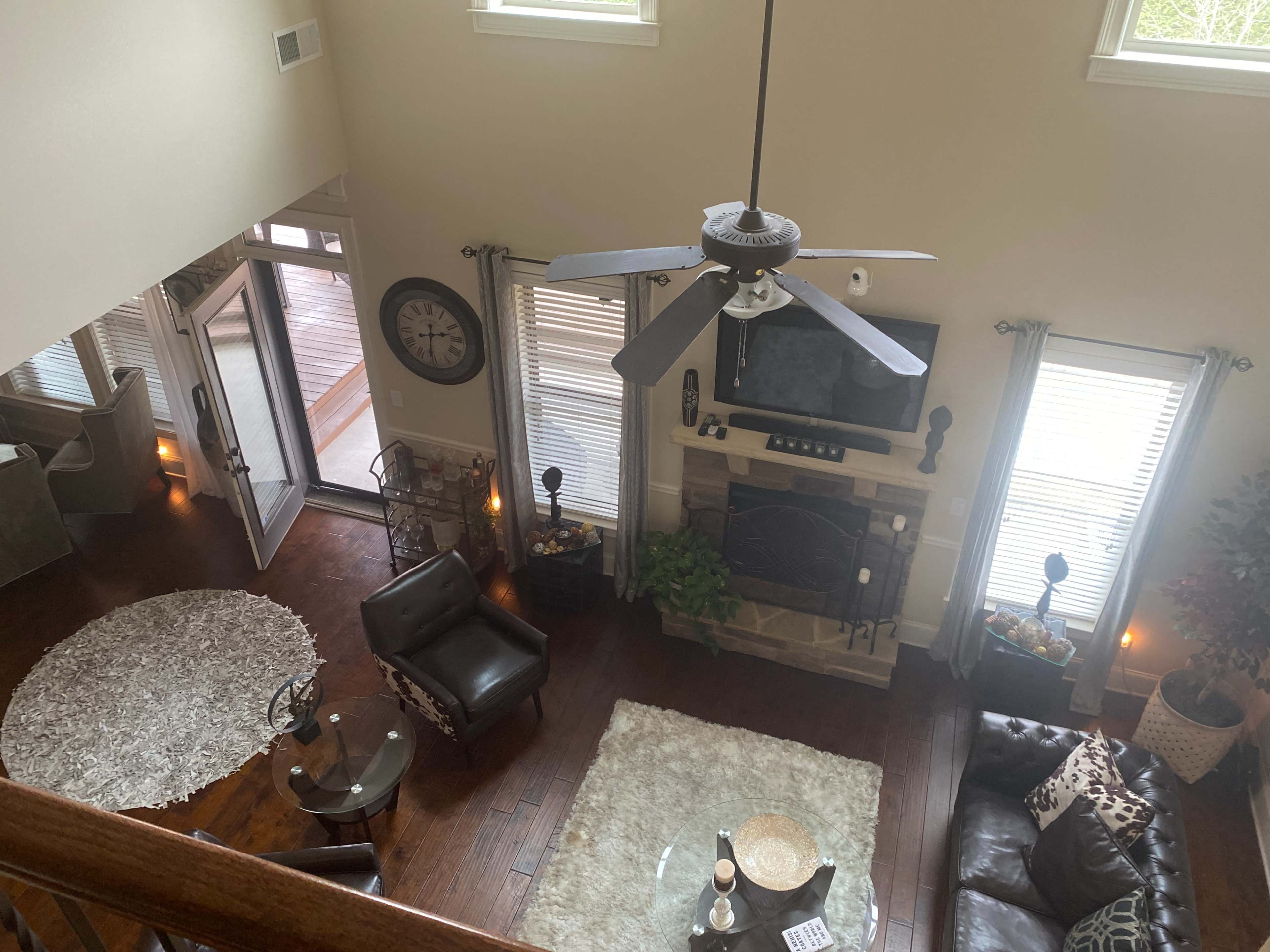 A spacious living room with a ceiling fan, two leather chairs, a coffee table, a rug, a fireplace, and large windows allowing natural light.