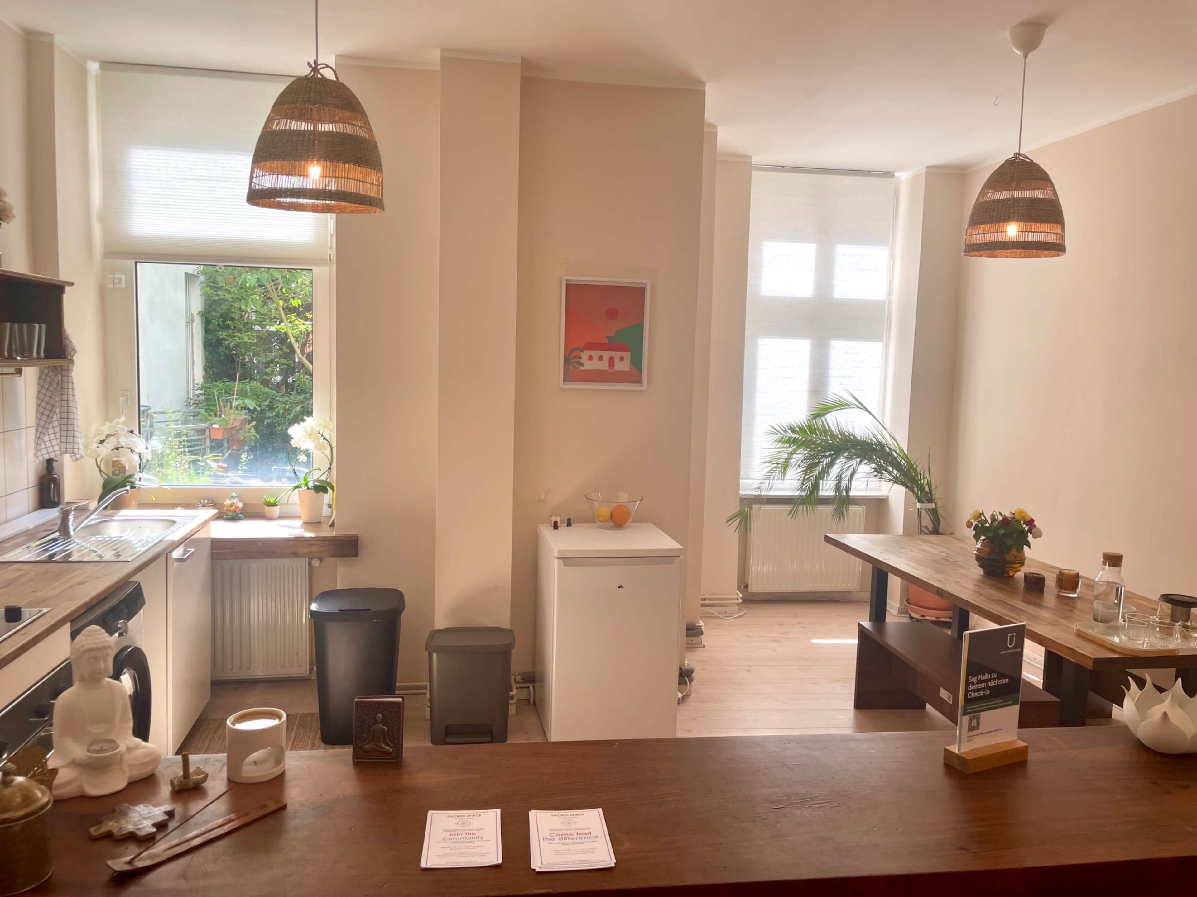 The image shows a bright kitchen area with wooden counters, a white refrigerator, and a window overlooking greenery, illuminated by pendant lights.