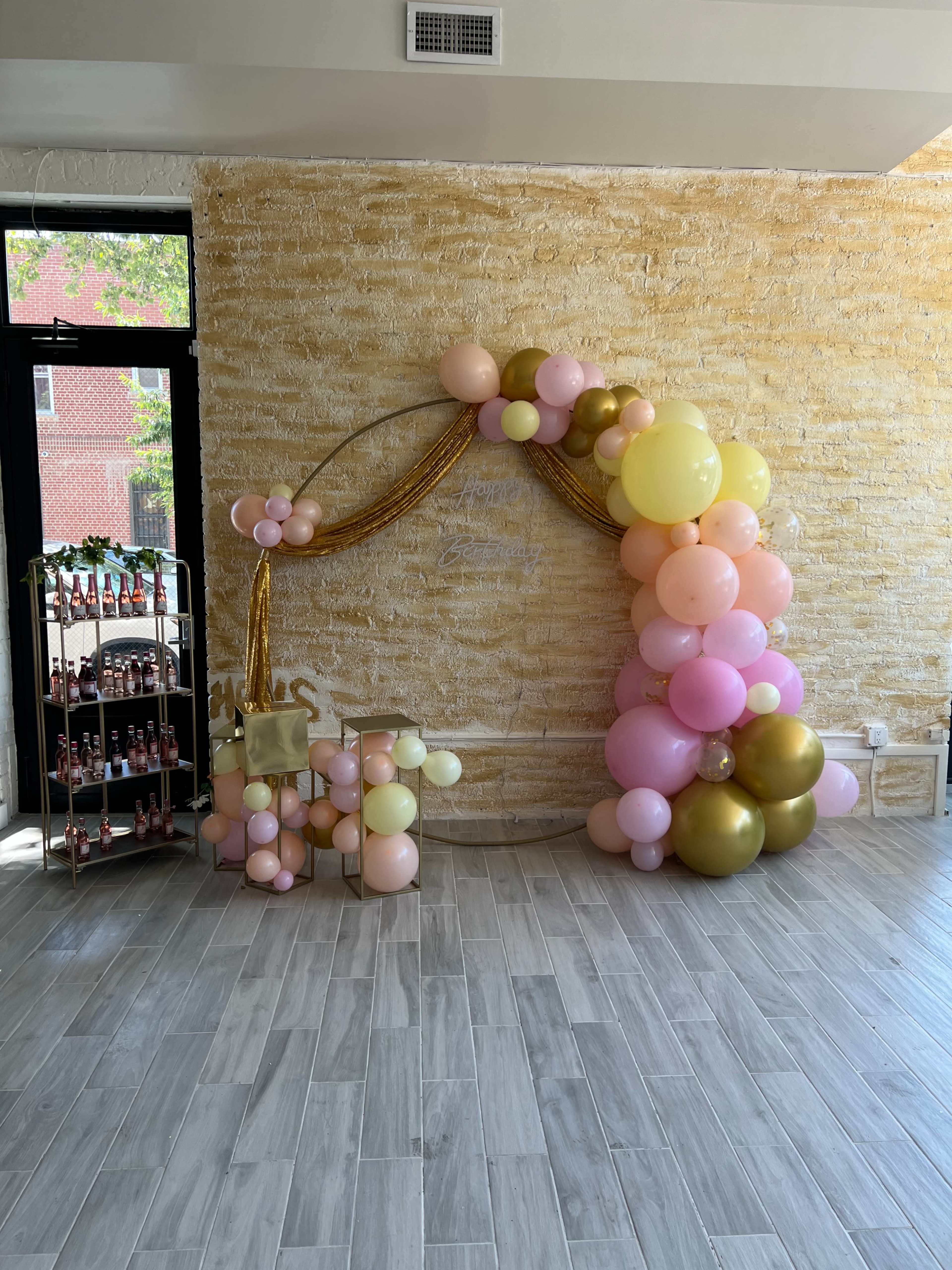 The image shows a decorated arch made of pink, gold, and yellow balloons against a textured light-colored wall, with a beverage cart in the background.