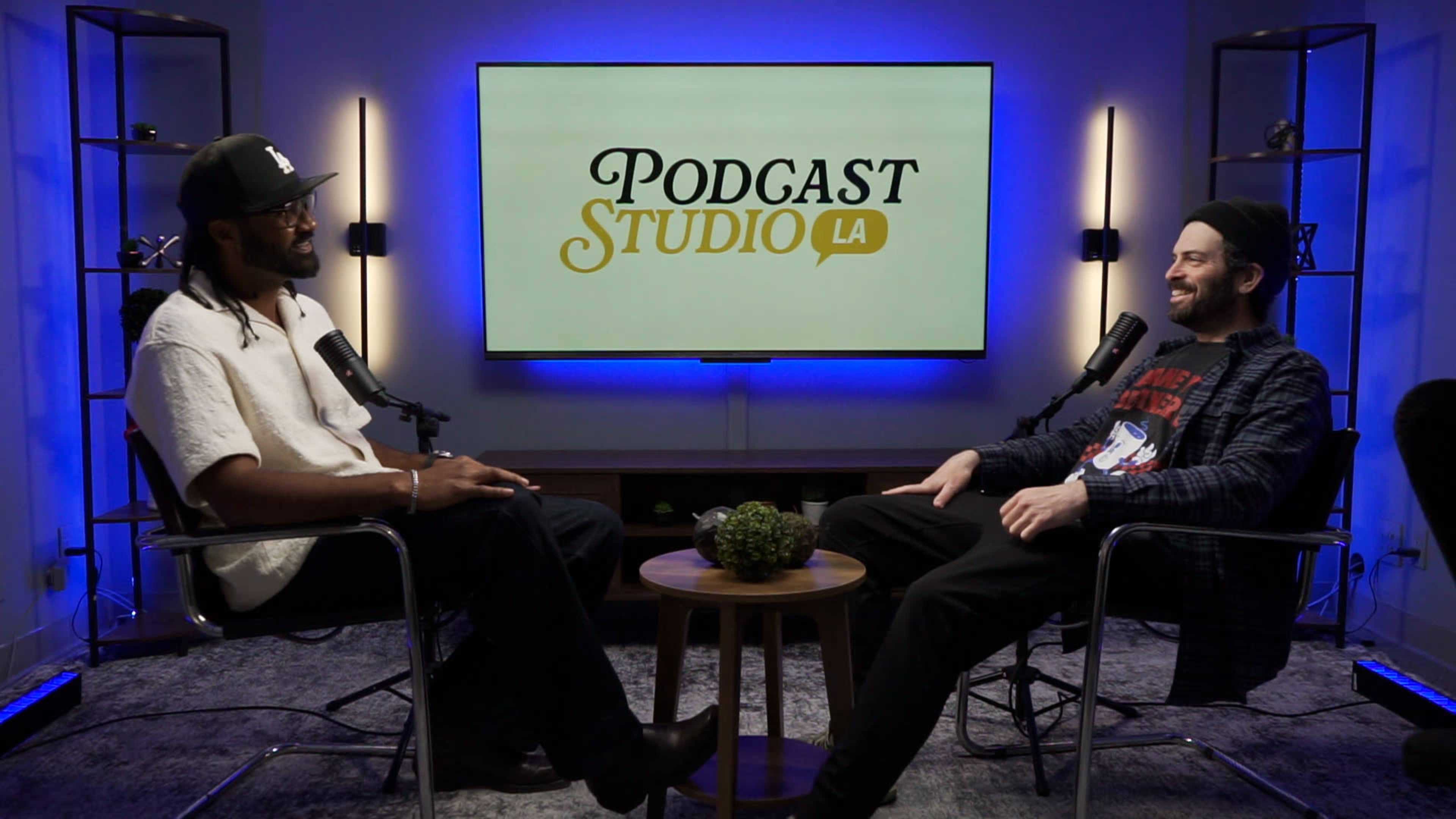 Two hosts sit in chairs across from each other in a podcast studio, with a large screen displaying "Podcast Studio LA" behind them.