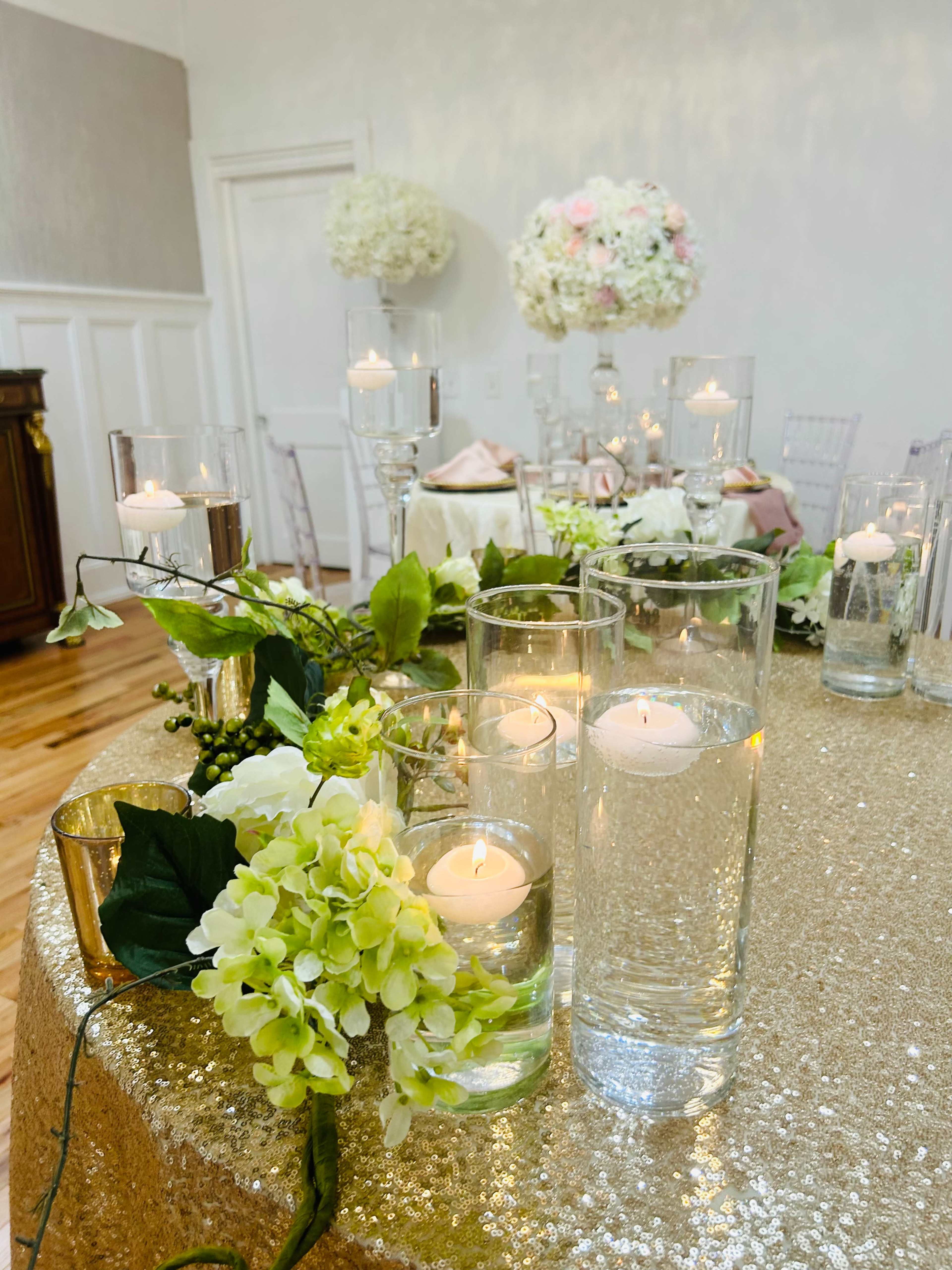 The image shows a elegantly arranged table with floral centerpieces, candles in glass containers, and a sequined tablecloth.