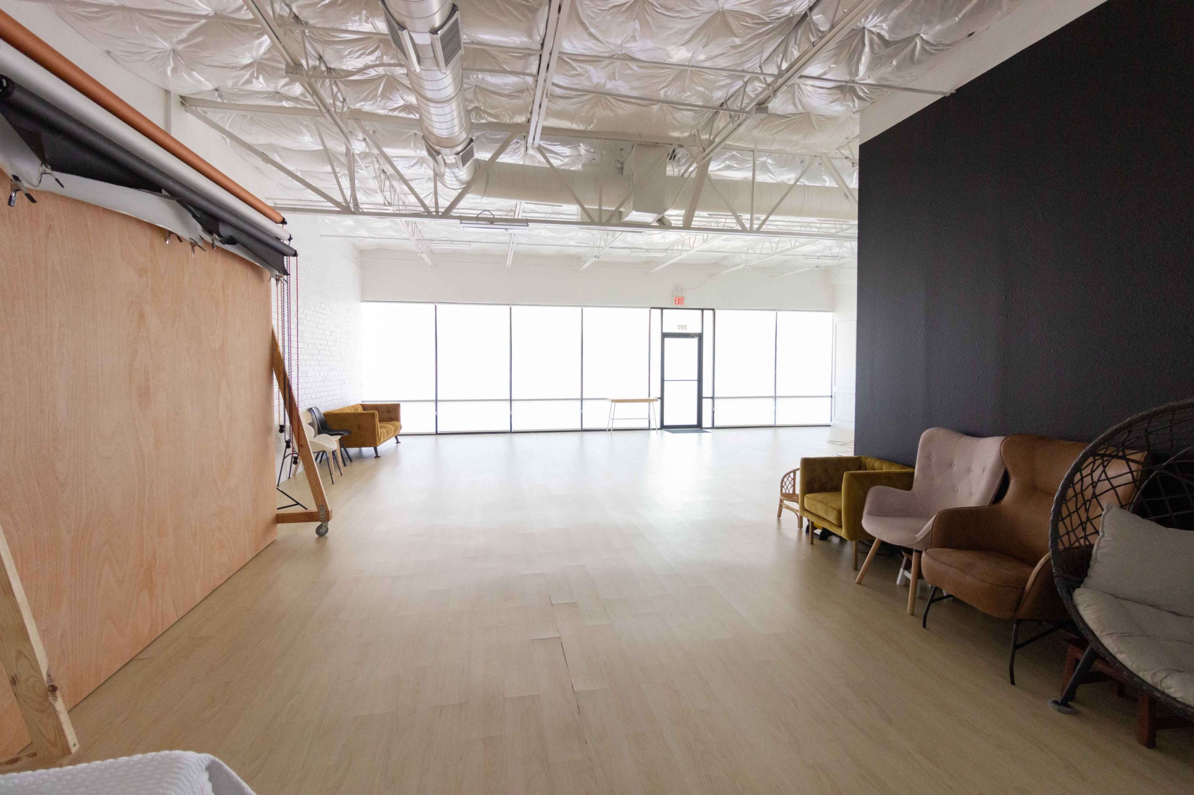 A spacious, well-lit studio with large windows, wooden flooring, and a few distinct seating areas including chairs and a wooden backdrop.