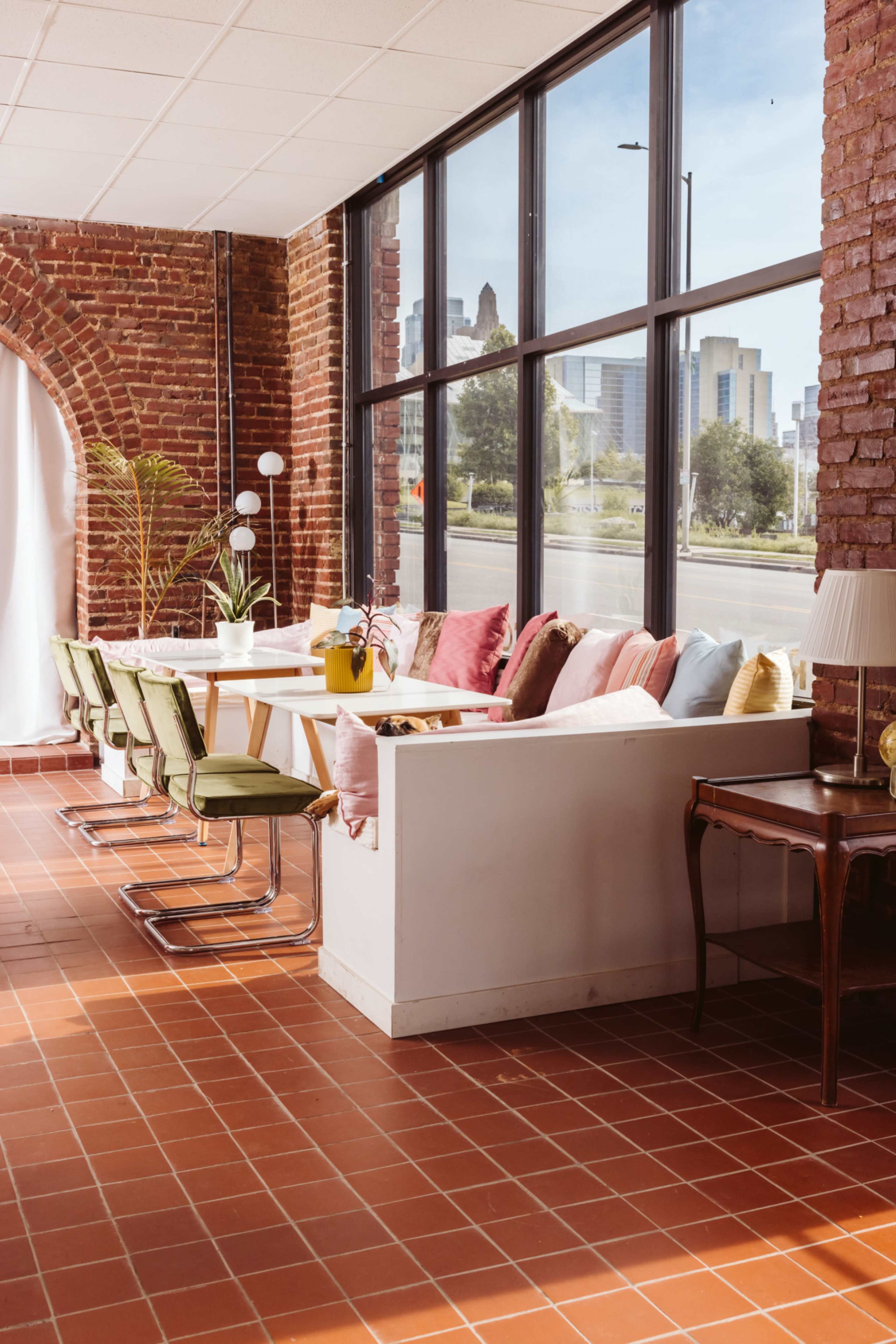 A bright interior features a long table with colorful cushions along a brick wall, large windows providing a city view, and green chairs.