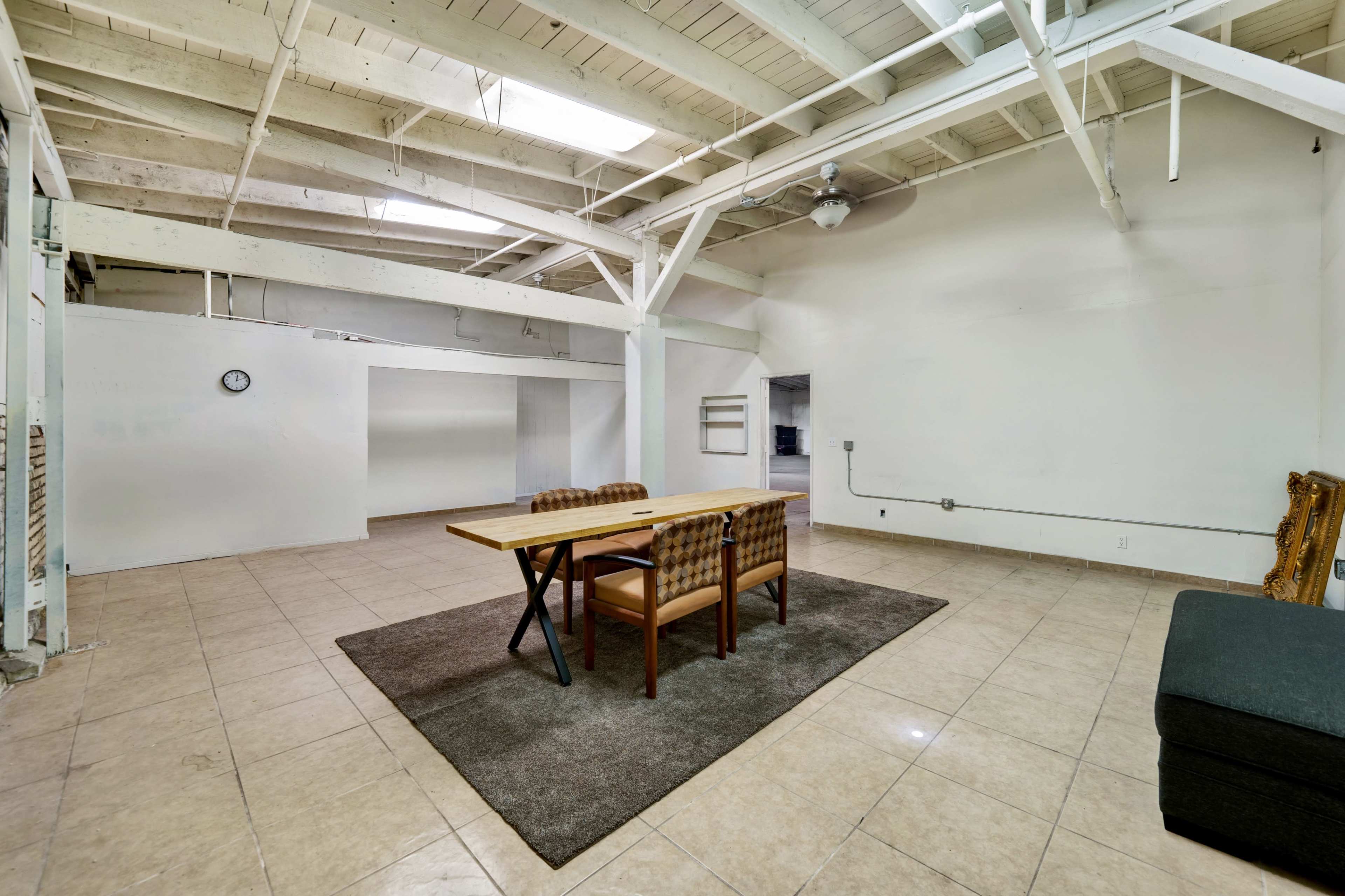 A spacious room with a wooden dining table and chairs on a carpet, surrounded by white walls and a high ceiling.
