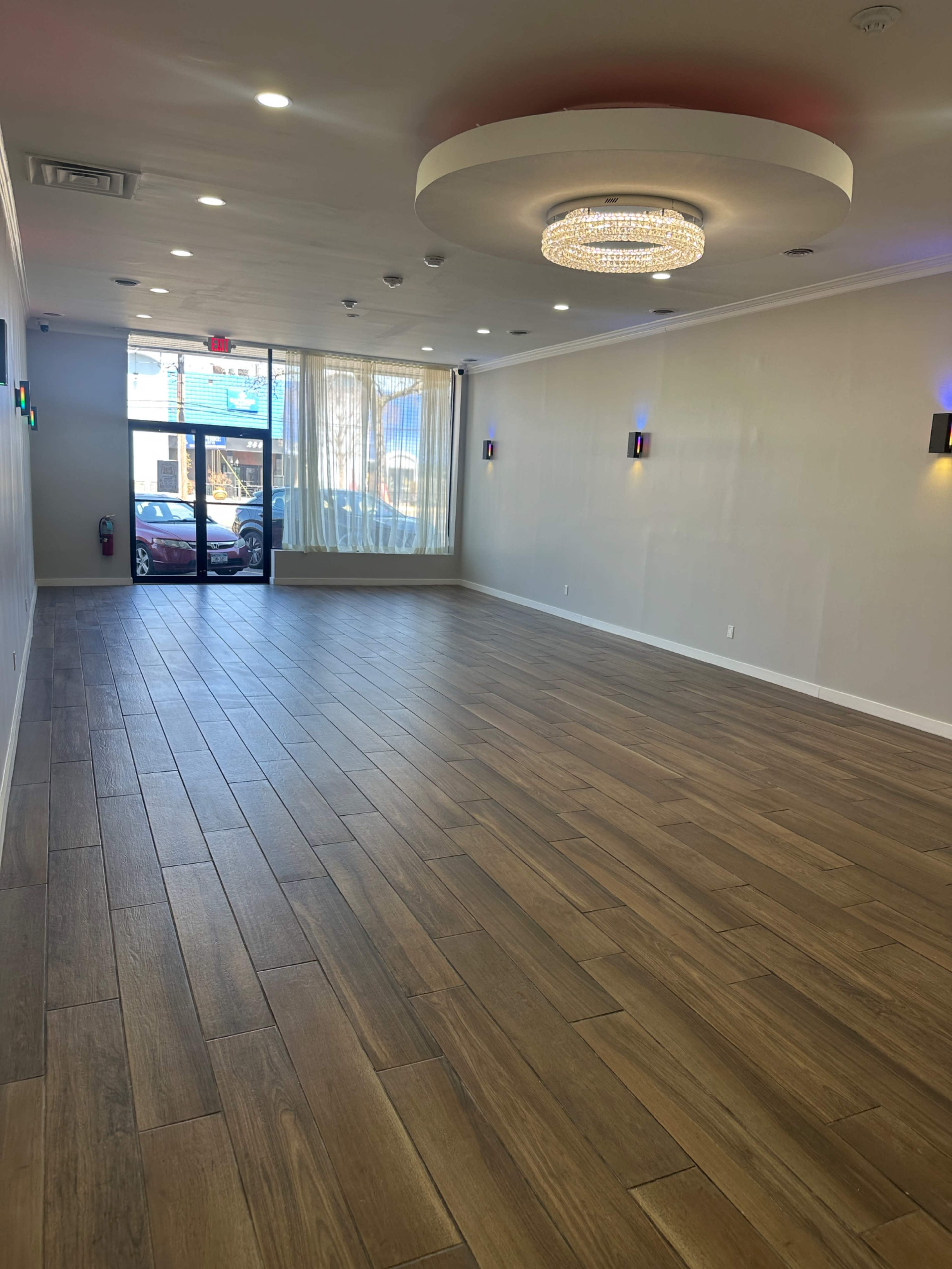 The image shows an empty, well-lit interior space with wooden flooring, large windows allowing natural light, and a modern chandelier overhead.