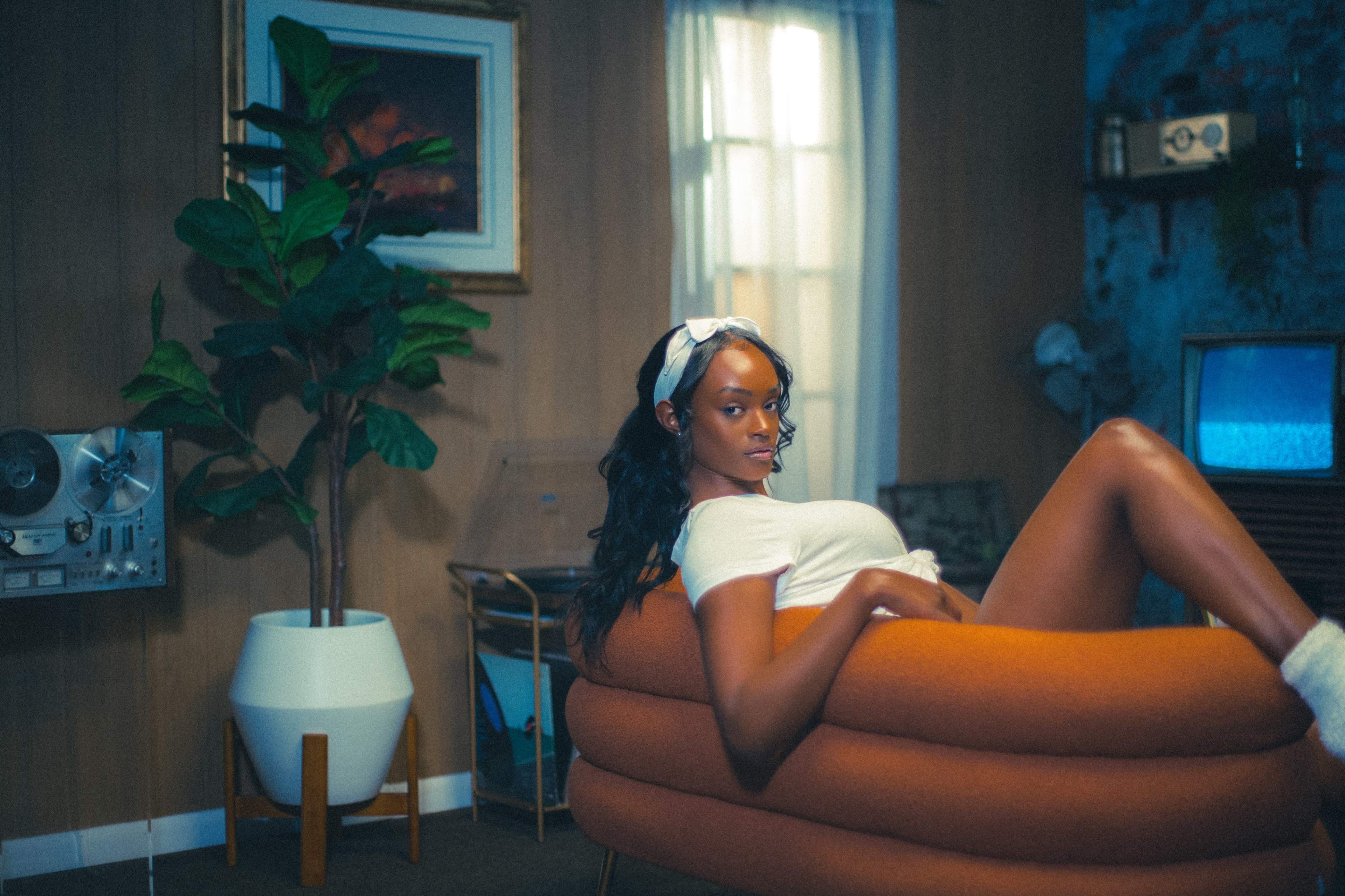 A woman sits comfortably on an orange, curved chair in a room with vintage decor, including plants and a retro television.