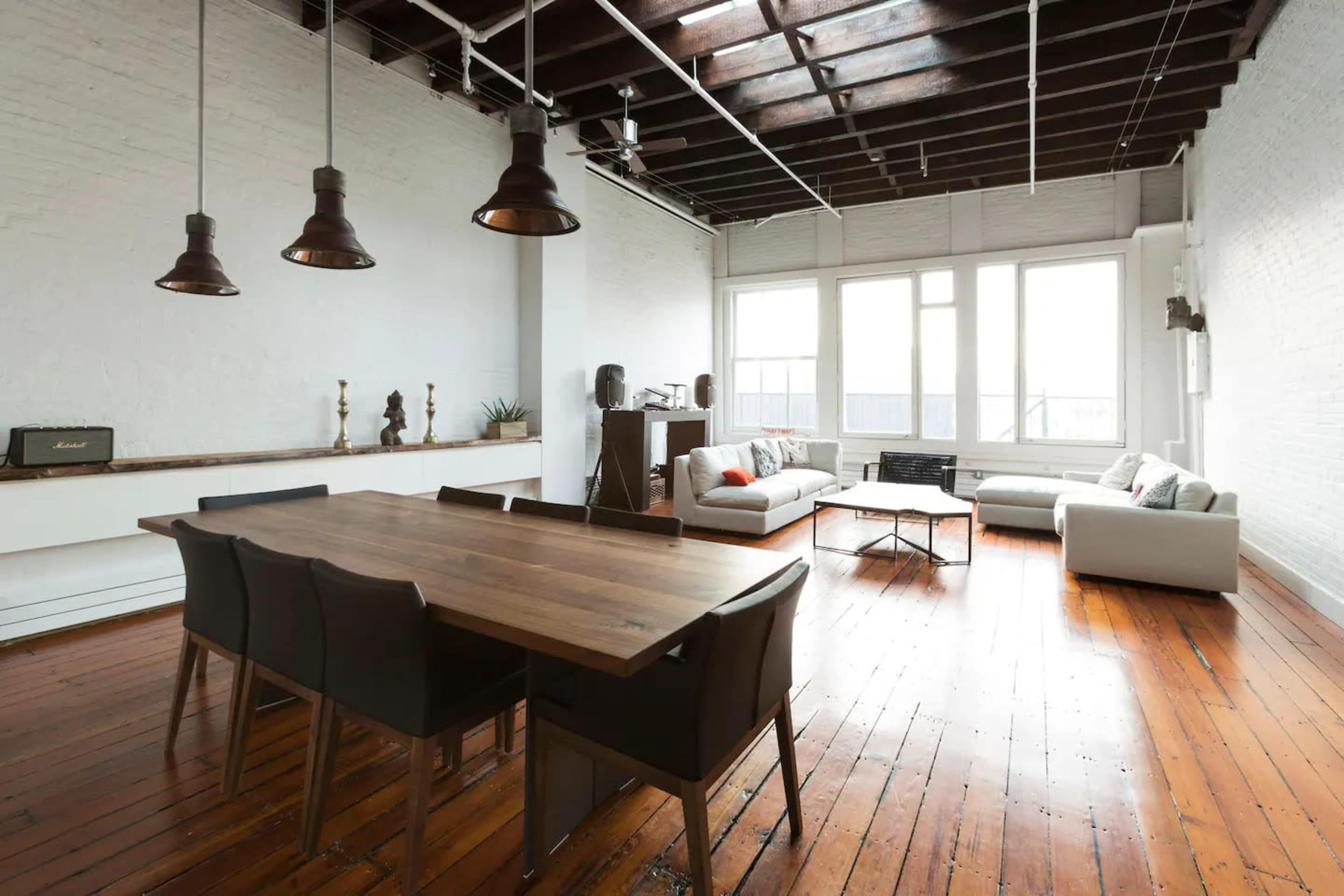 A spacious, well-lit interior featuring a dining table with chairs, a seating area with sofas, and large windows allowing natural light to enter.
