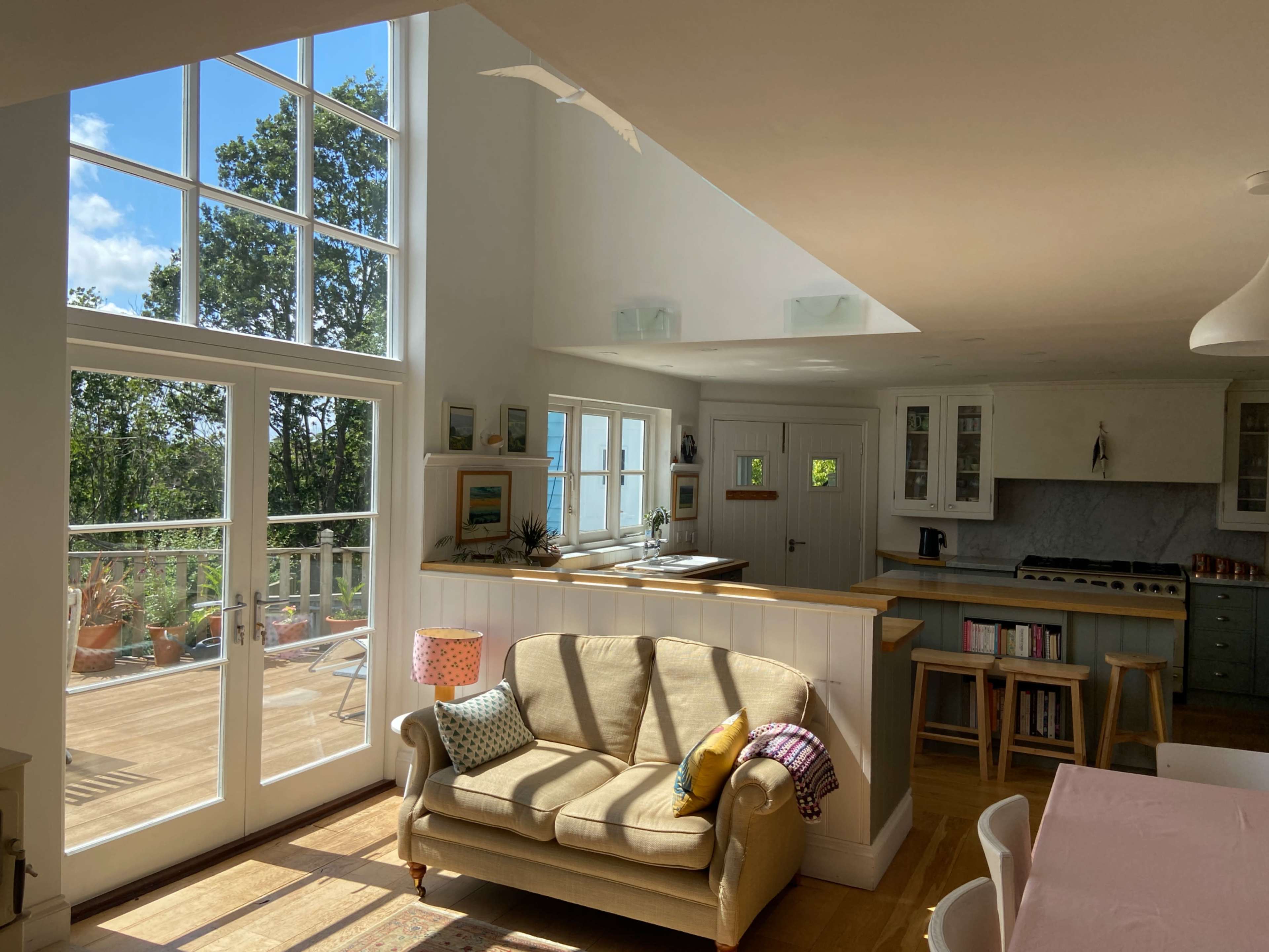 The image shows a bright, modern living space featuring a sofa, large windows, and access to a deck with potted plants.