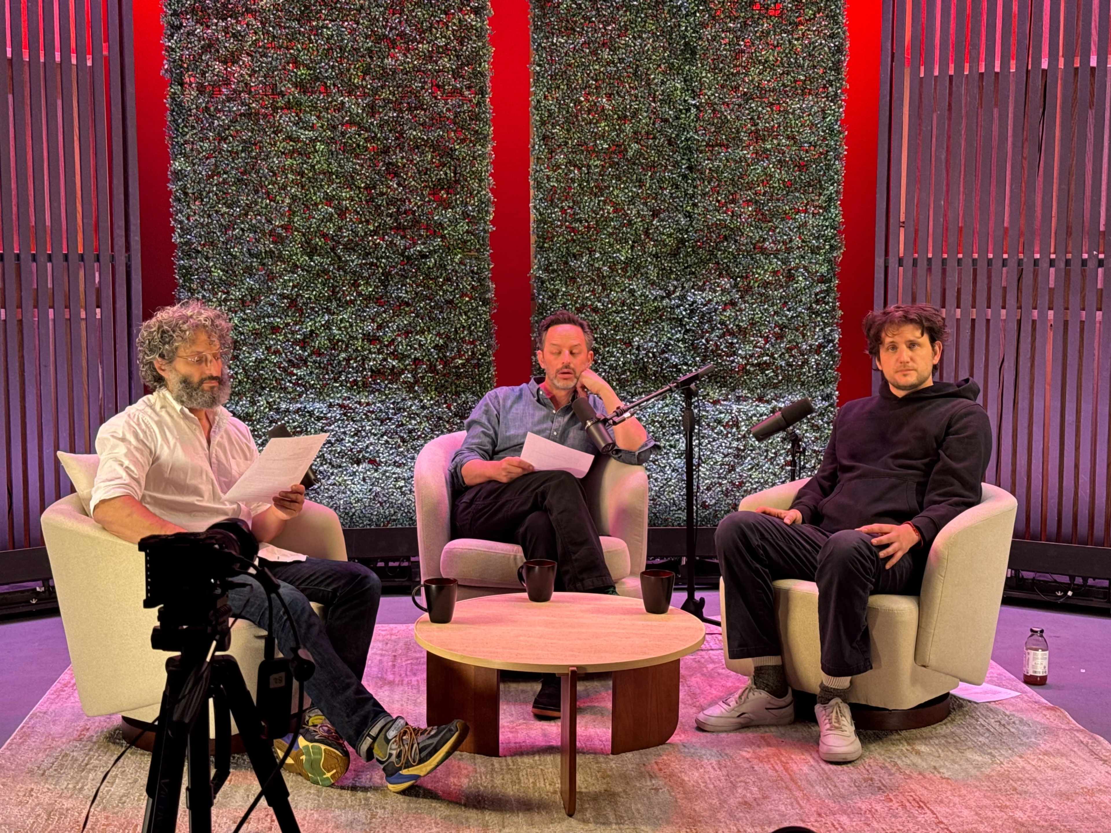Three men sit in a studio setting with a wooden table and microphones, surrounded by a green wall, as they review their notes.