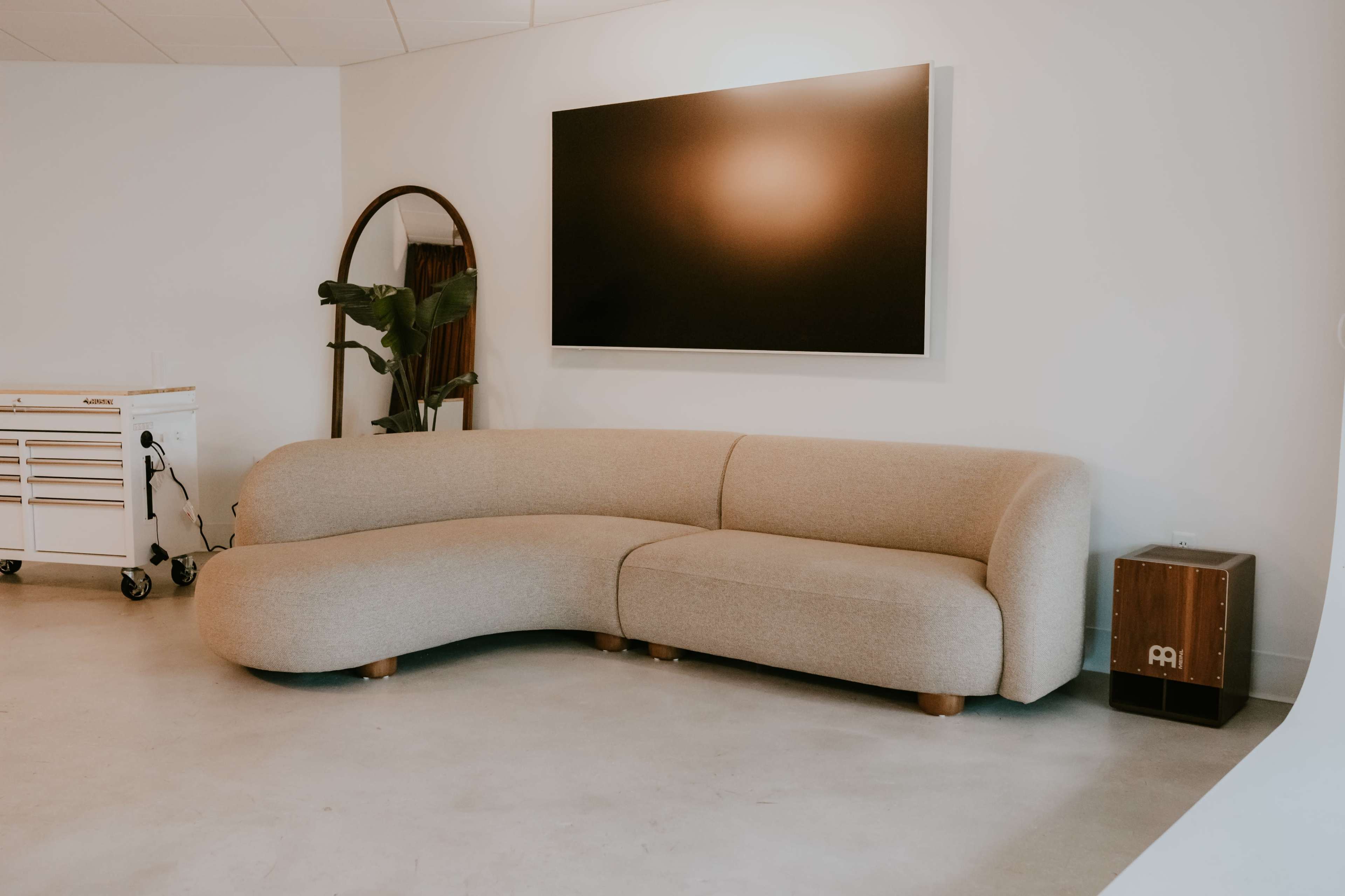 The scene features a modern, curved beige sofa positioned against a white wall, accompanied by a flat-screen television mounted above it and a small wooden table nearby.