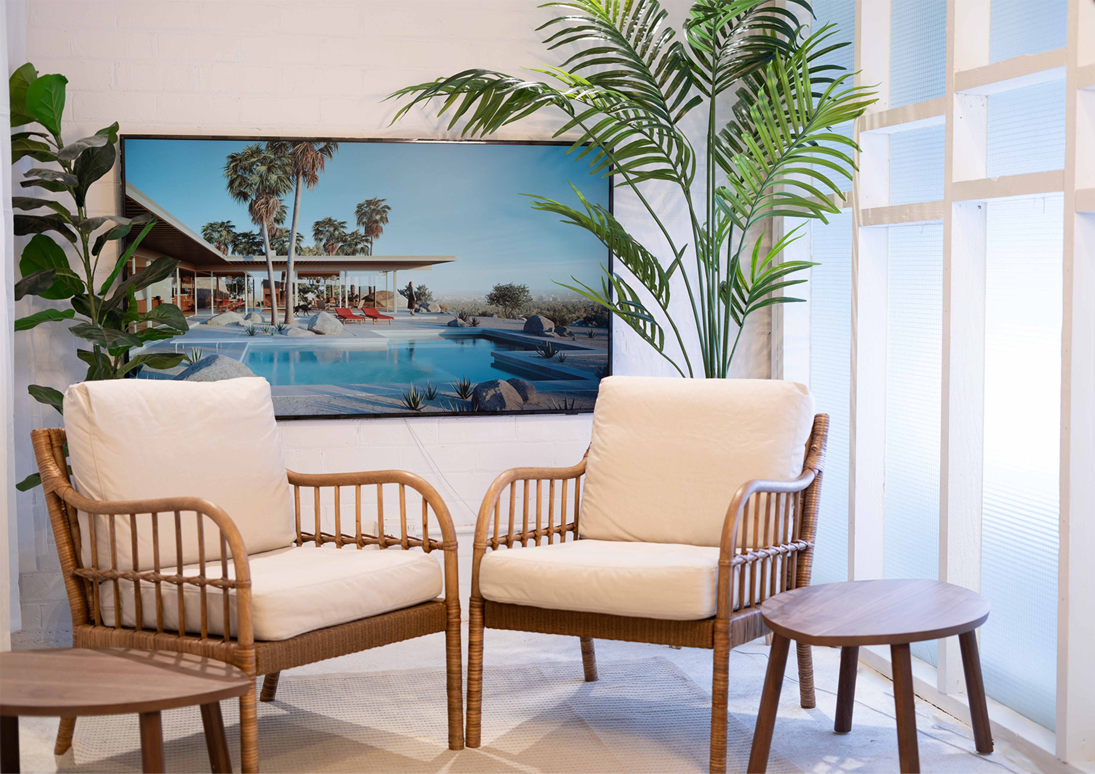 Two wicker armchairs with cream cushions, a small round wooden table, and a large framed landscape photograph of a modern poolside scene with palm trees.