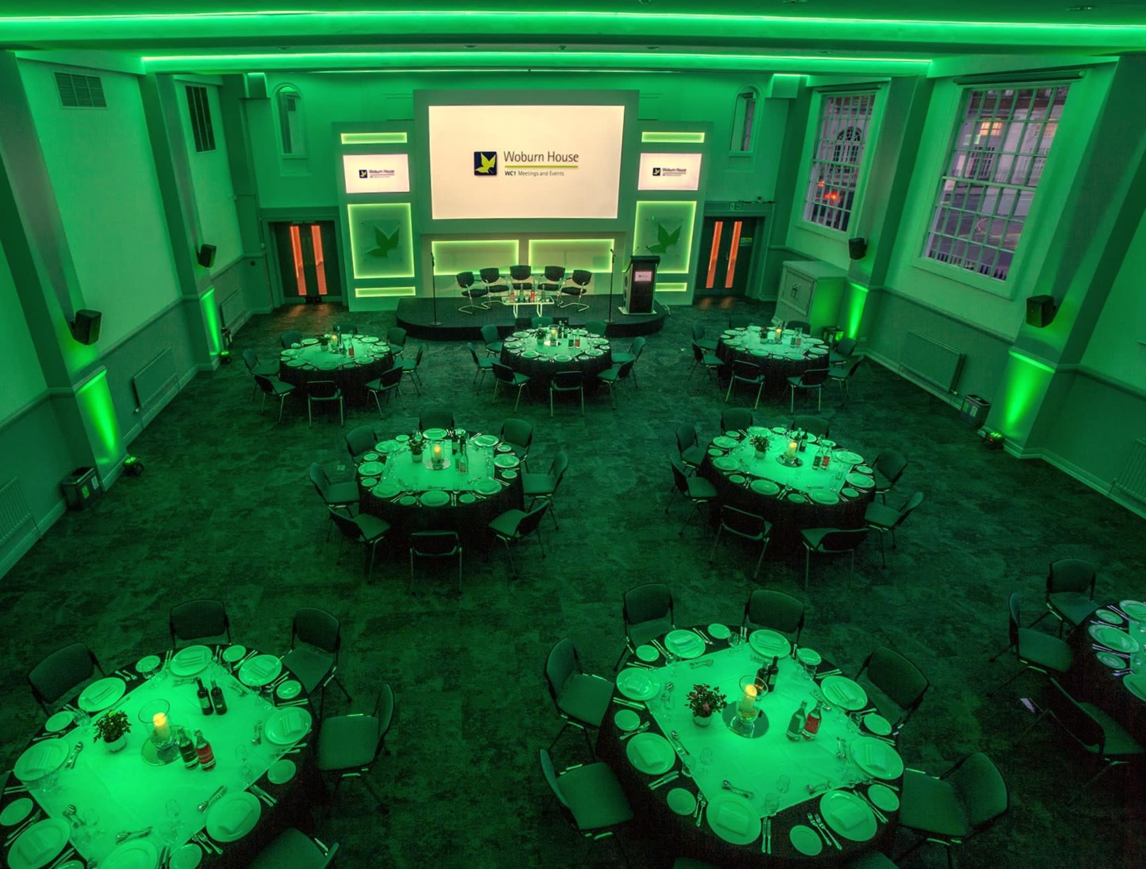 A banquet hall with round tables set for dining under green ambient lighting, featuring a presentation screen at the front.