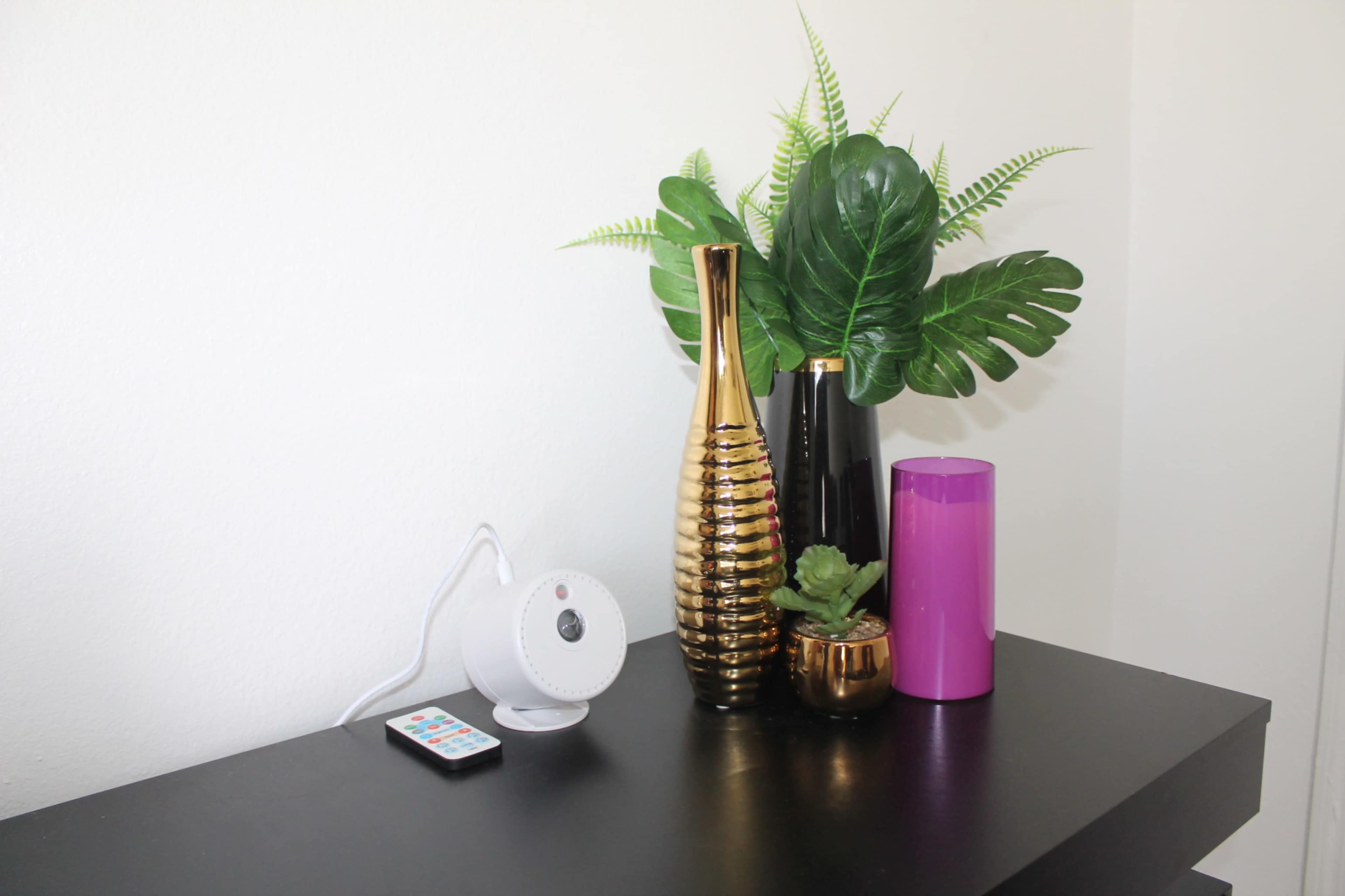 A decorative arrangement features a gold vase, a black vase, a pink cylindrical container, a small plant, and a remote on a black surface against a light-colored wall.
