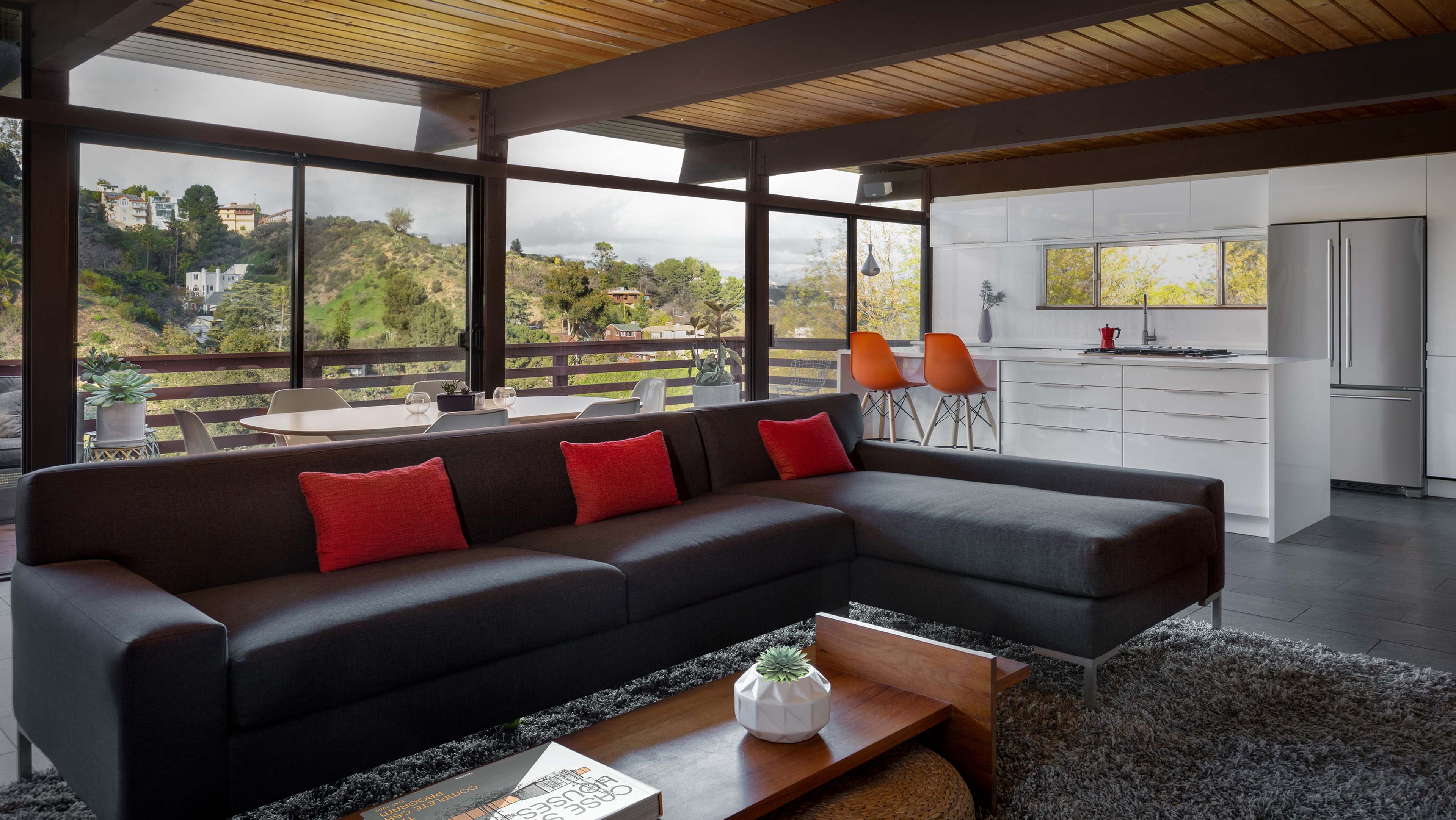 A modern living room features a dark sectional sofa with red accent pillows, large windows showcasing a hillside view, and a kitchen area with sleek white cabinetry.