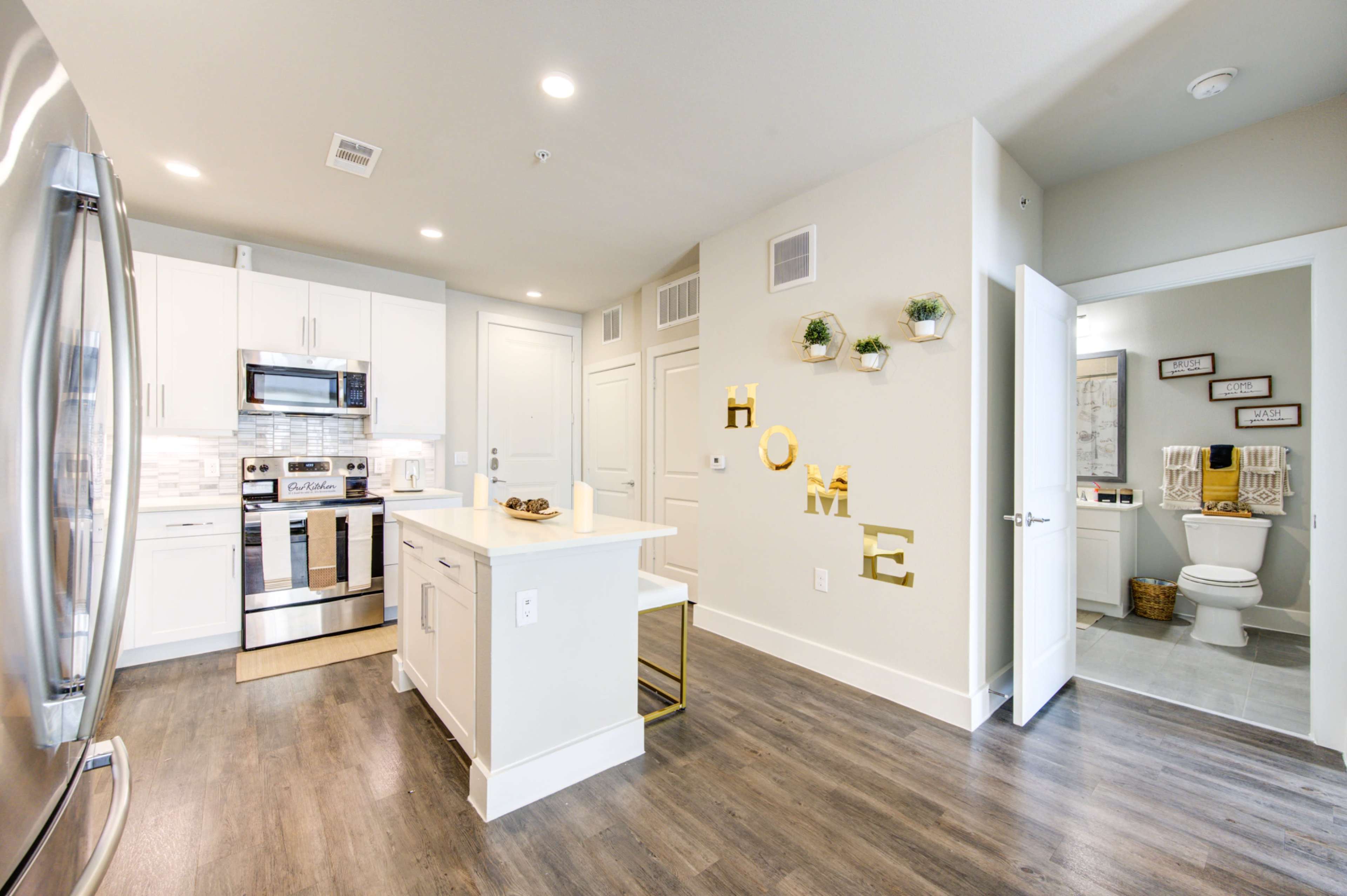 The image shows a modern kitchen with stainless steel appliances, white cabinets, and a small island, adjacent to a bathroom and decorated with a "HOME" wall display and plants.
