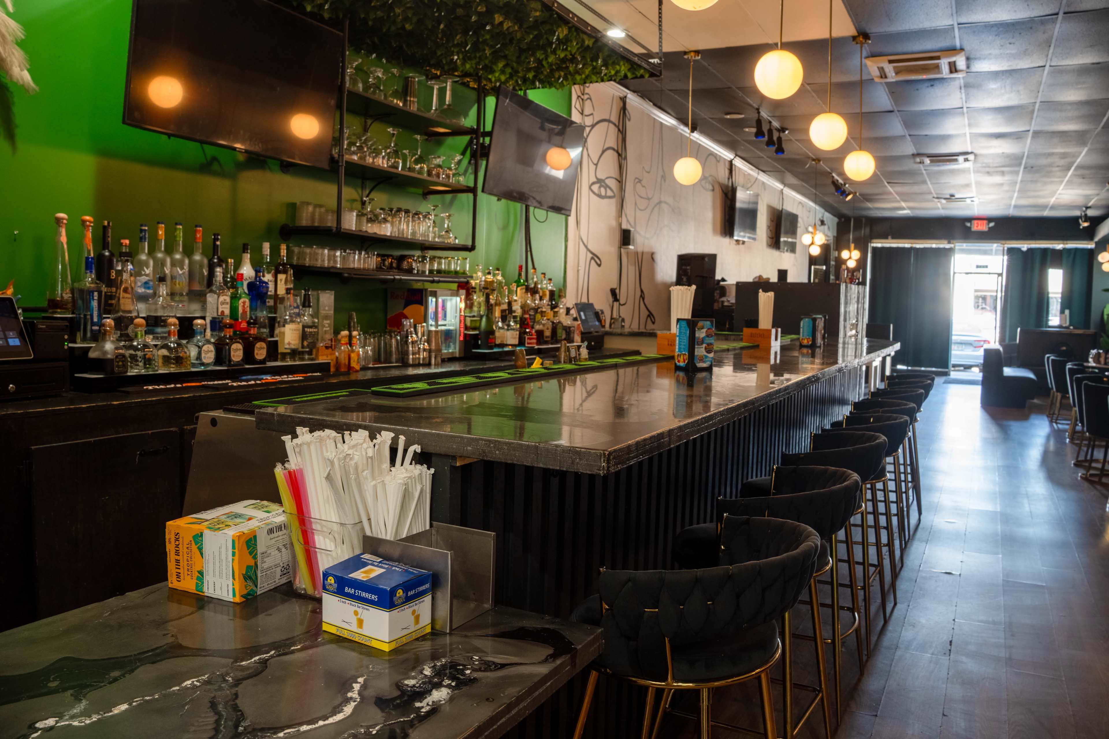 The image shows a modern bar interior with a long countertop, shelves of liquor bottles, and stylish seating along the bar.