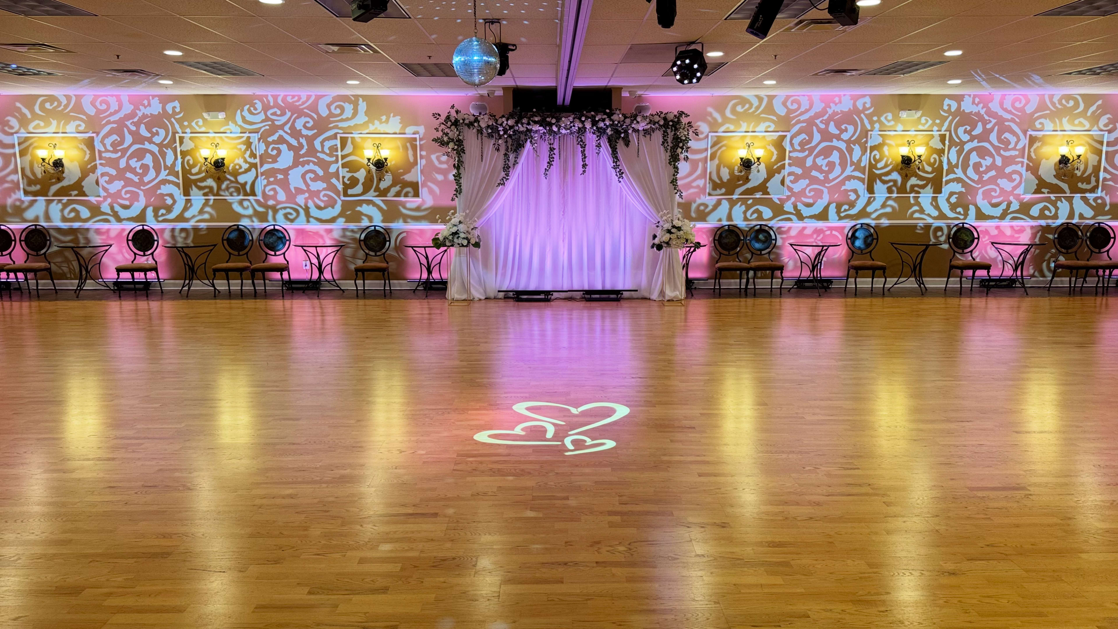 The image shows a spacious event hall with a wooden floor, decorated with pink lighting, a floral arch at the front, and heart projections on the floor.