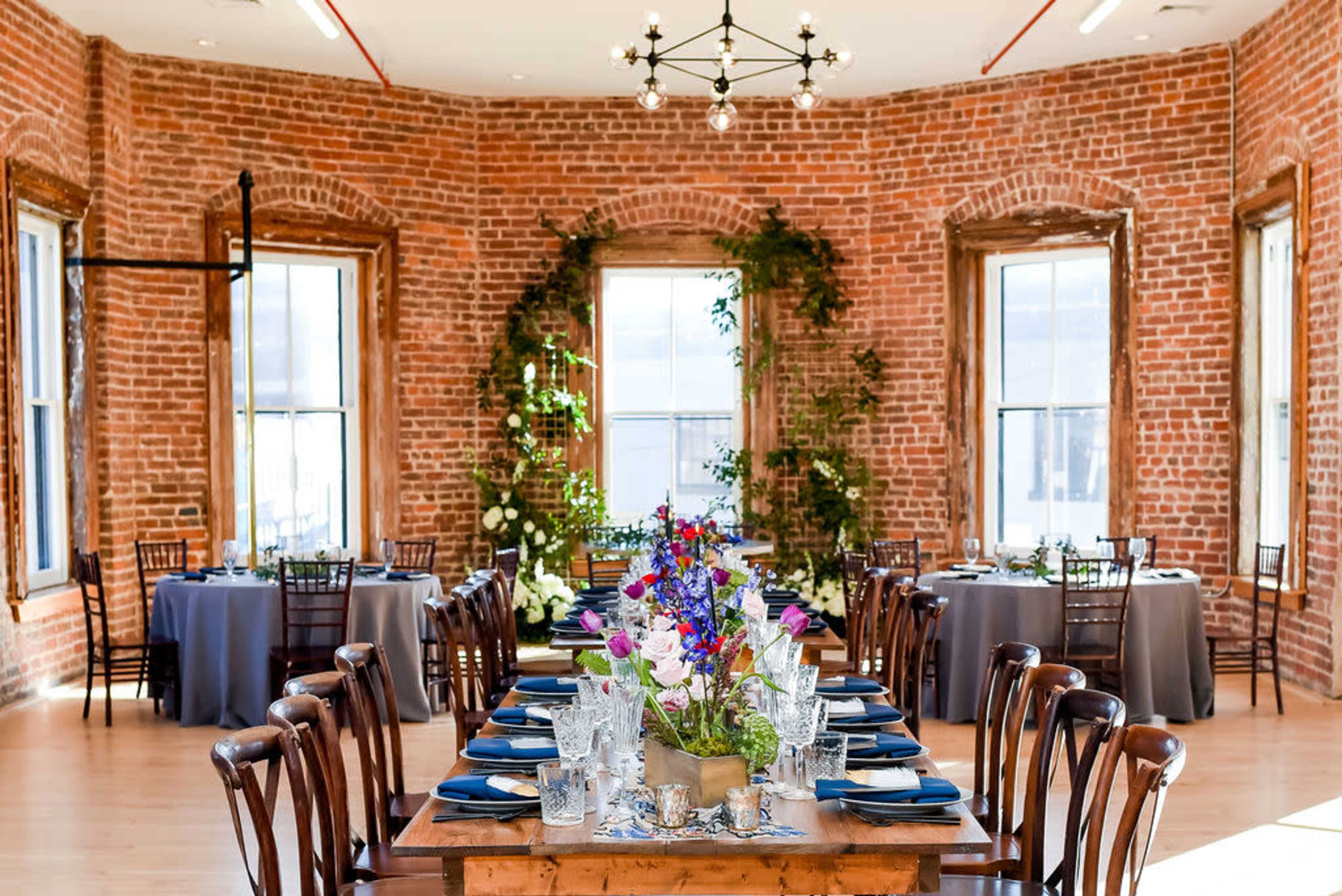 A banquet room with brick walls, large windows, and tables set with blue tableware and floral centerpieces.