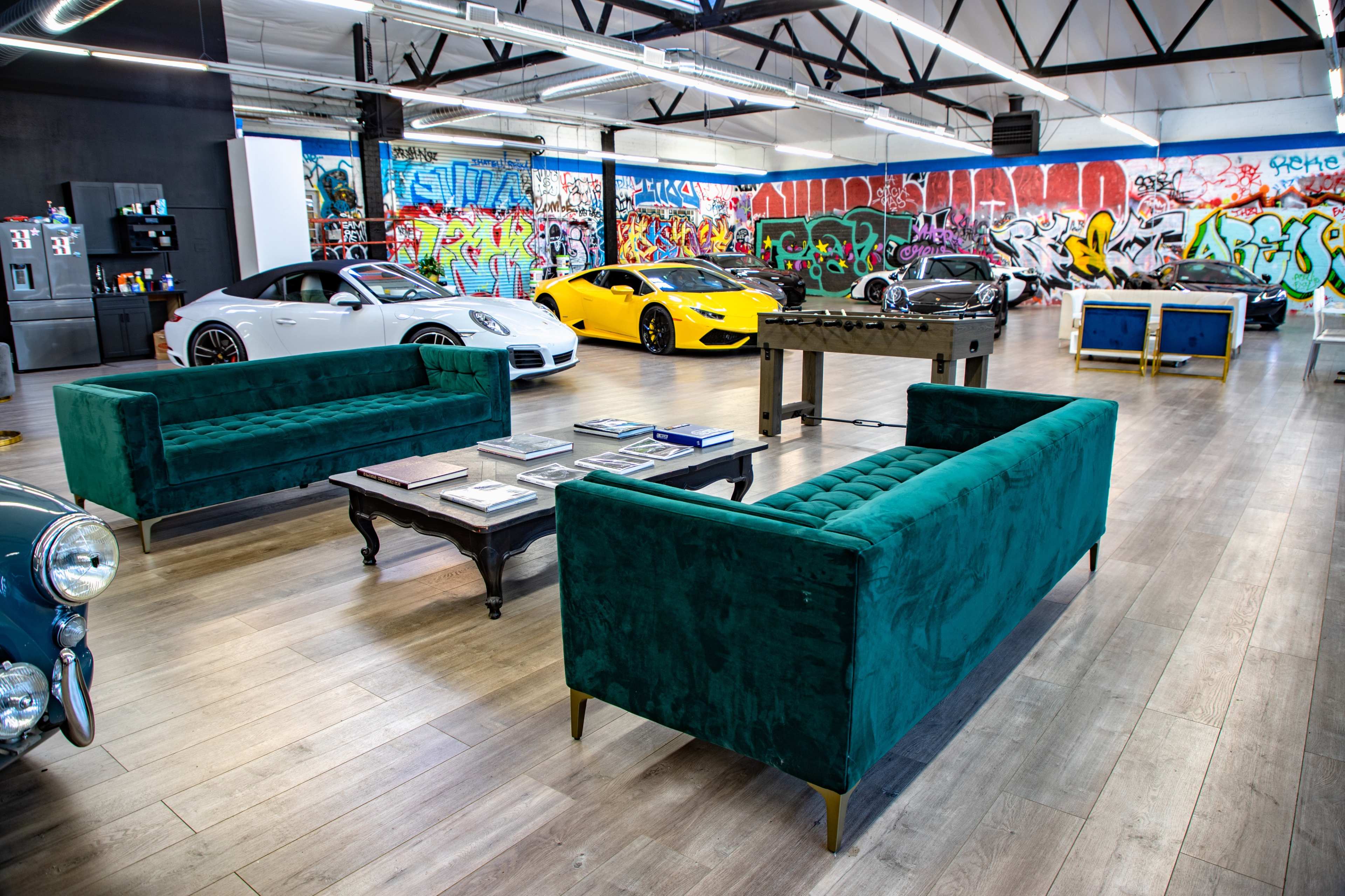 A spacious garage with modern cars, green velvet sofas, a coffee table with magazines, and vibrant graffiti on the walls.