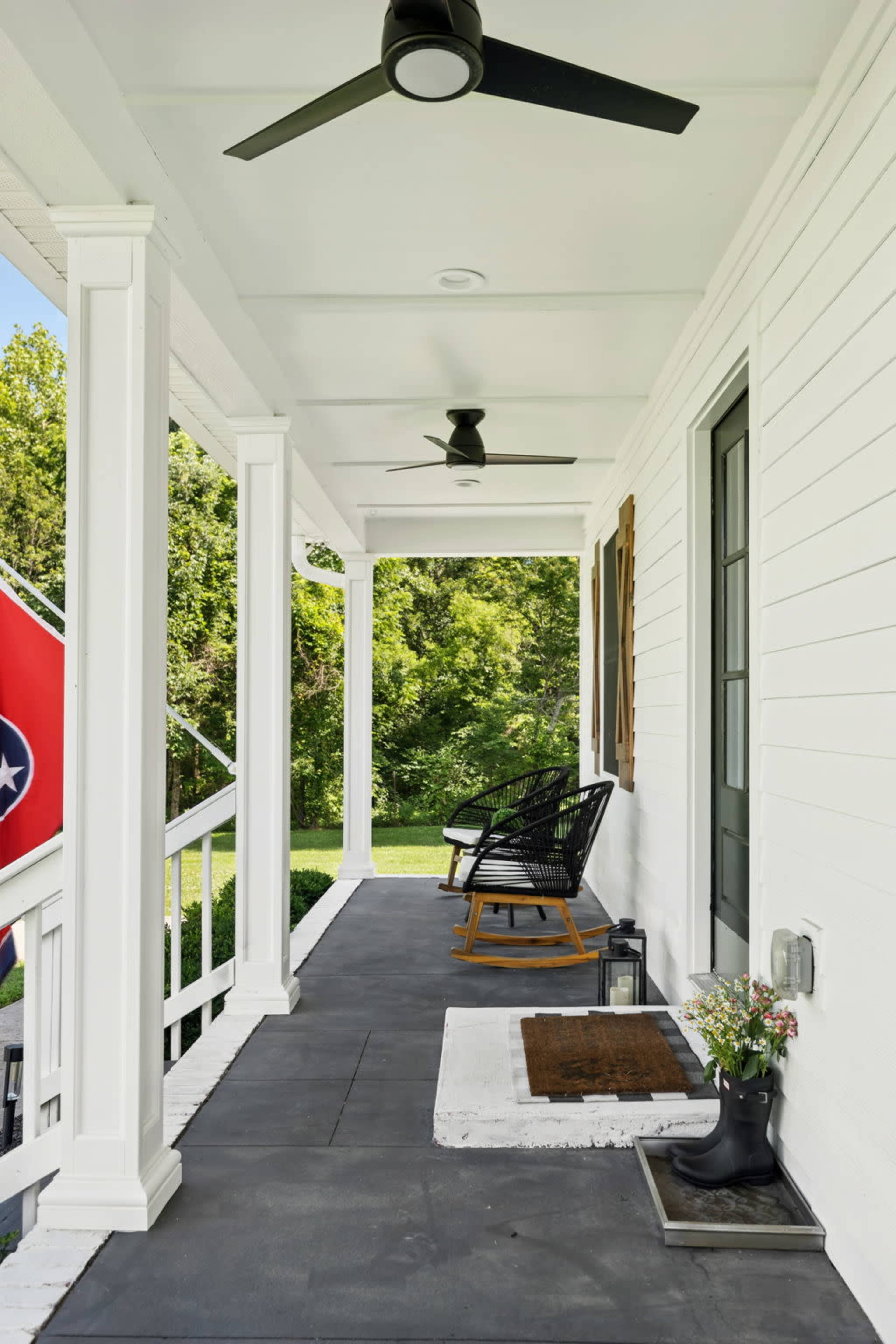 The image shows a porch with two ceiling fans, a black rocking chair, and a welcome mat next to a pair of black boots, framed by white columns and greenery in the background.