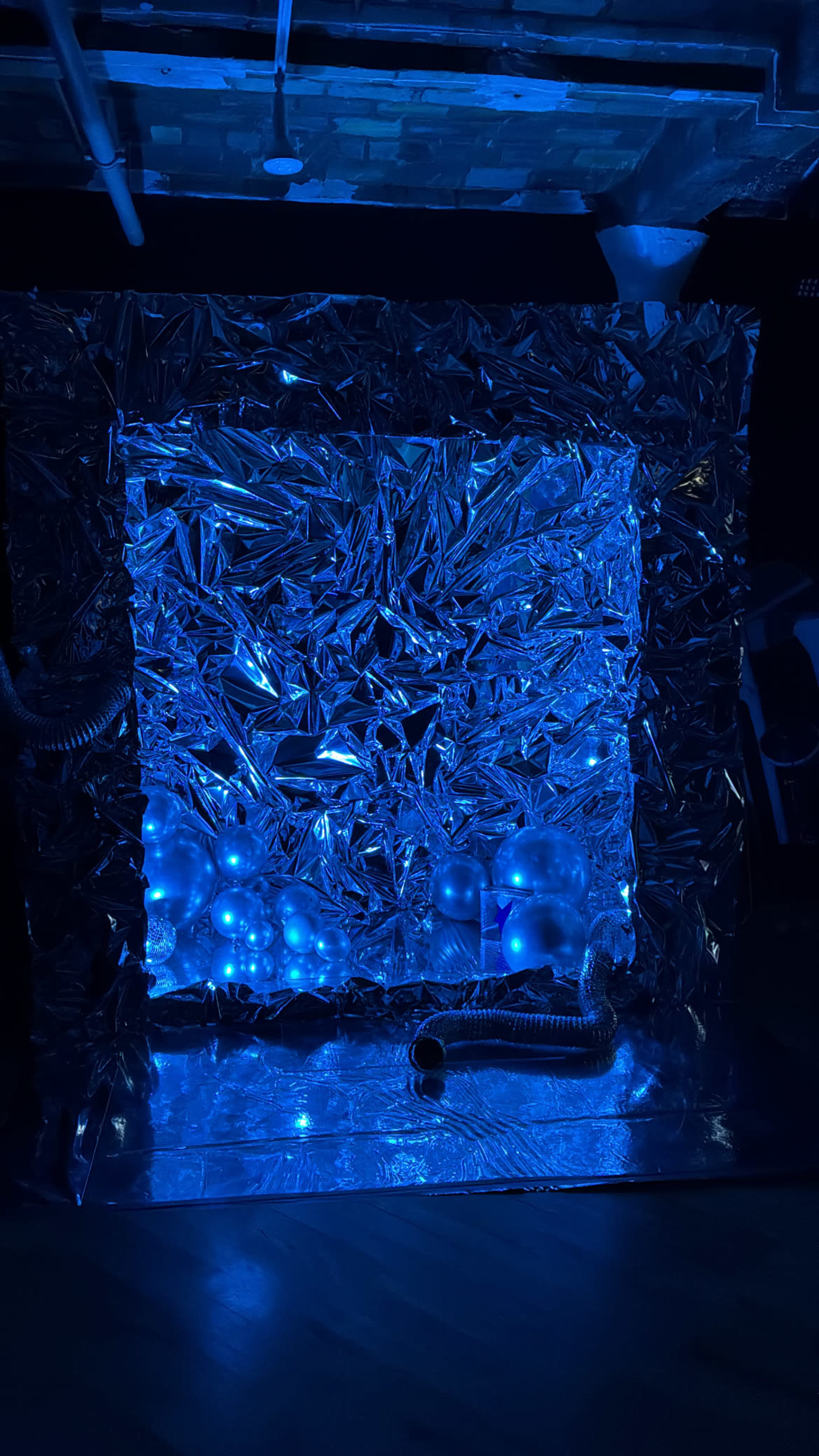 The image shows a blue-lit room featuring a reflective, crinkled silver backdrop with illuminated spheres and dark tubing at the base.