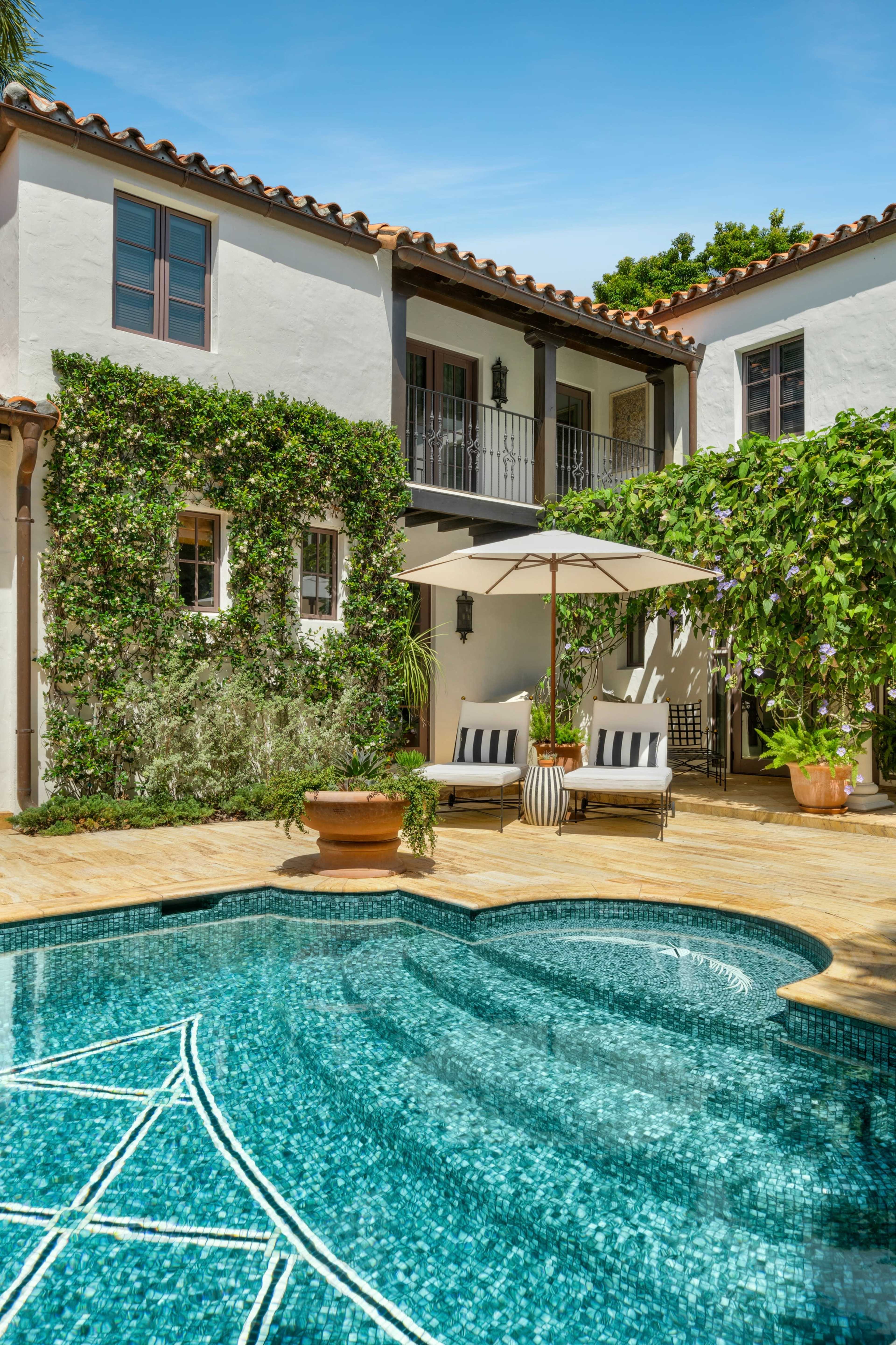 A swimming pool with a mosaic design is situated in a courtyard surrounded by a two-story building adorned with greenery and patio furniture.