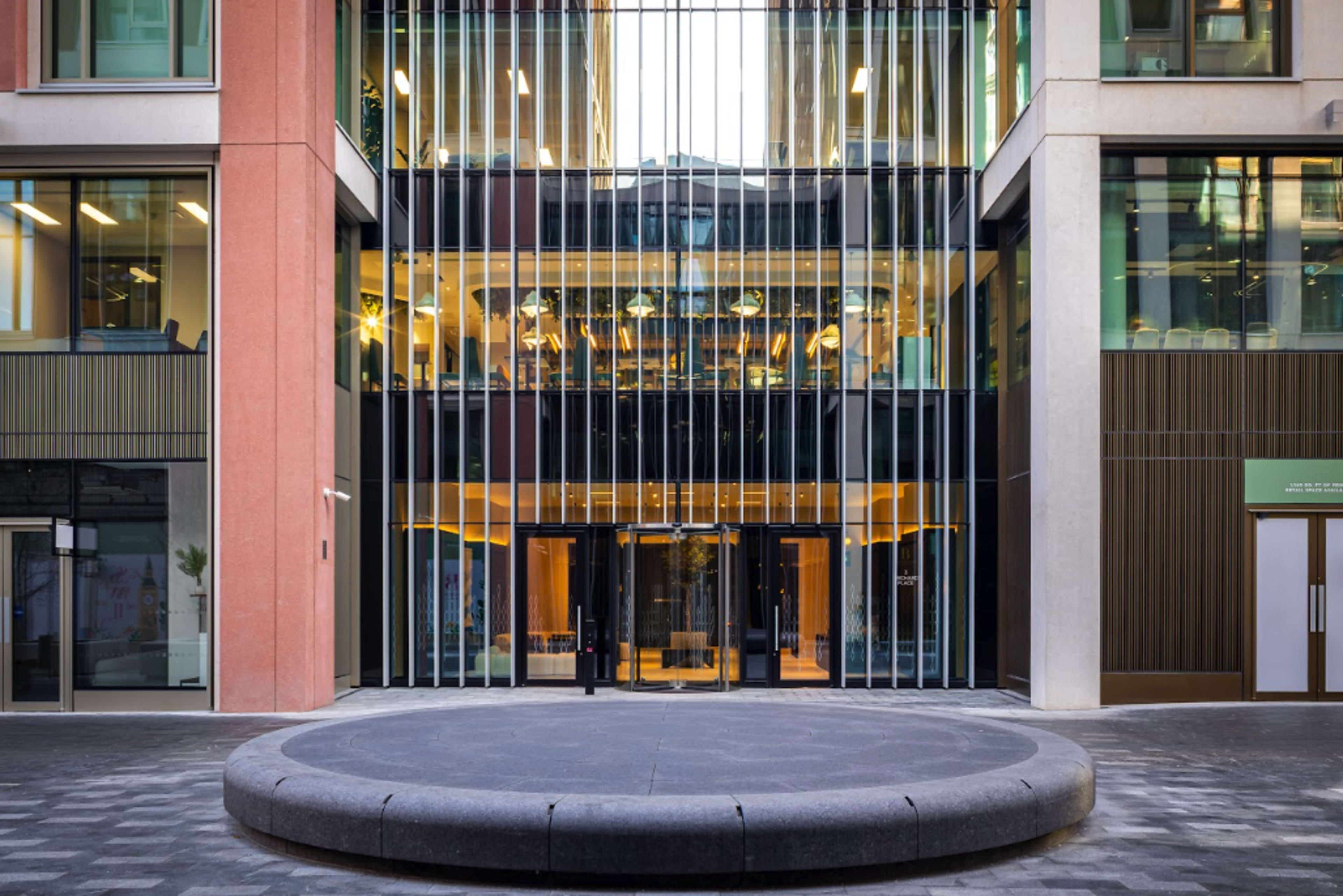 A modern building facade with glass walls and vertical strips of material, featuring a circular stone area at the entrance.