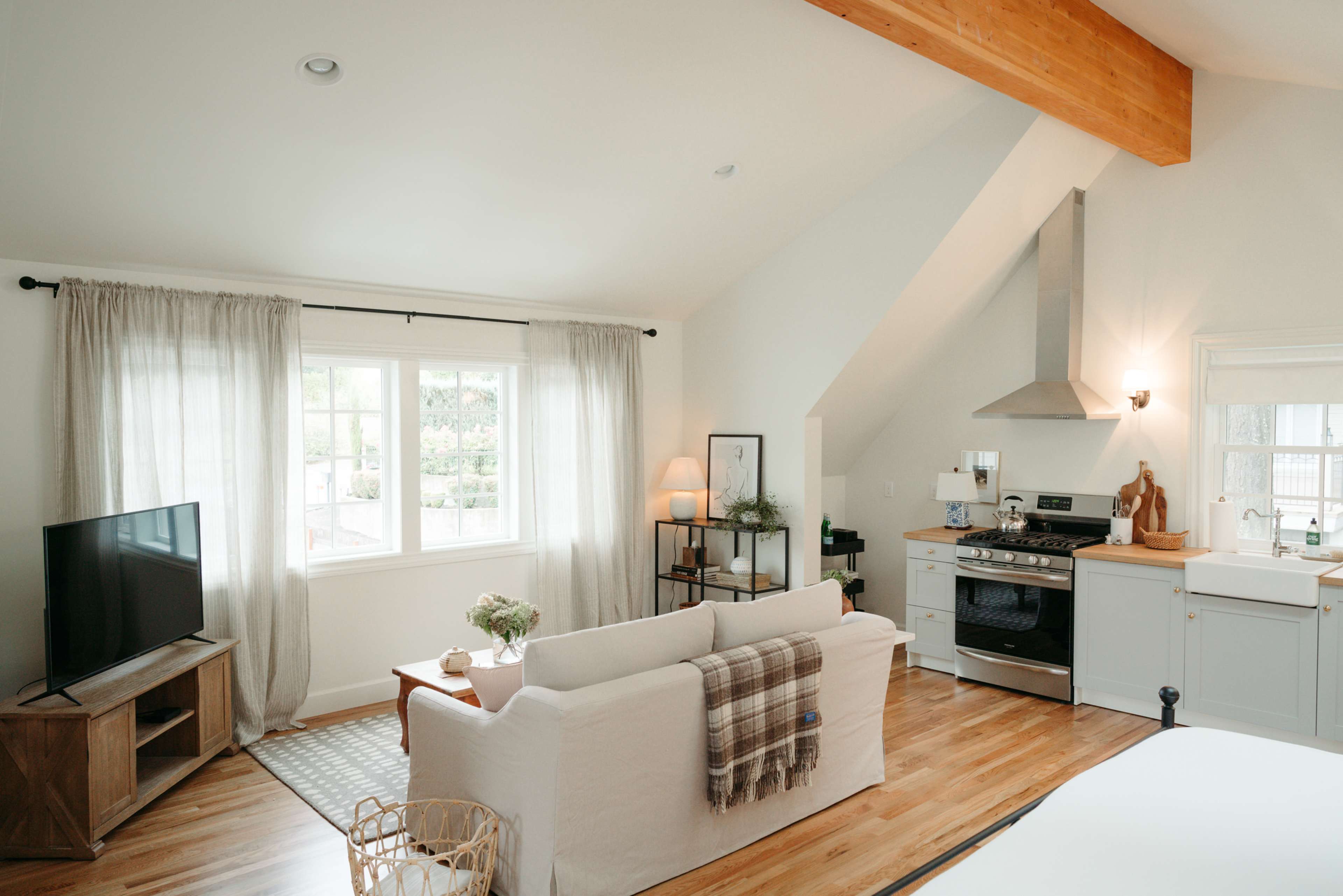 A well-lit open living space featuring a sofa with a blanket, a television, a kitchenette with a stove, and large windows with sheer curtains.