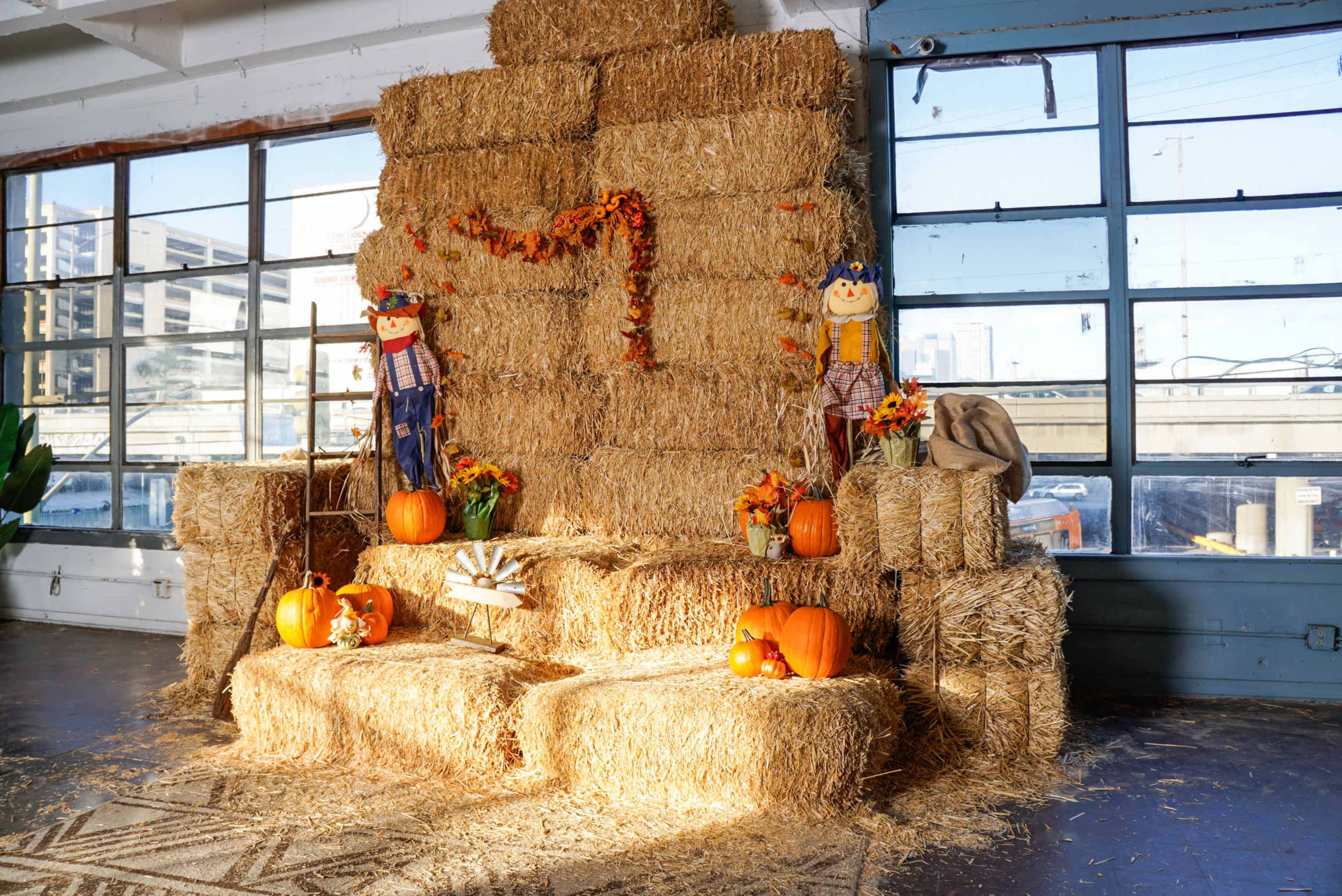 A decorative autumn scene featuring hay bales arranged in a backdrop, flanked by scarecrow figures, pumpkins, and fall-themed decorations.