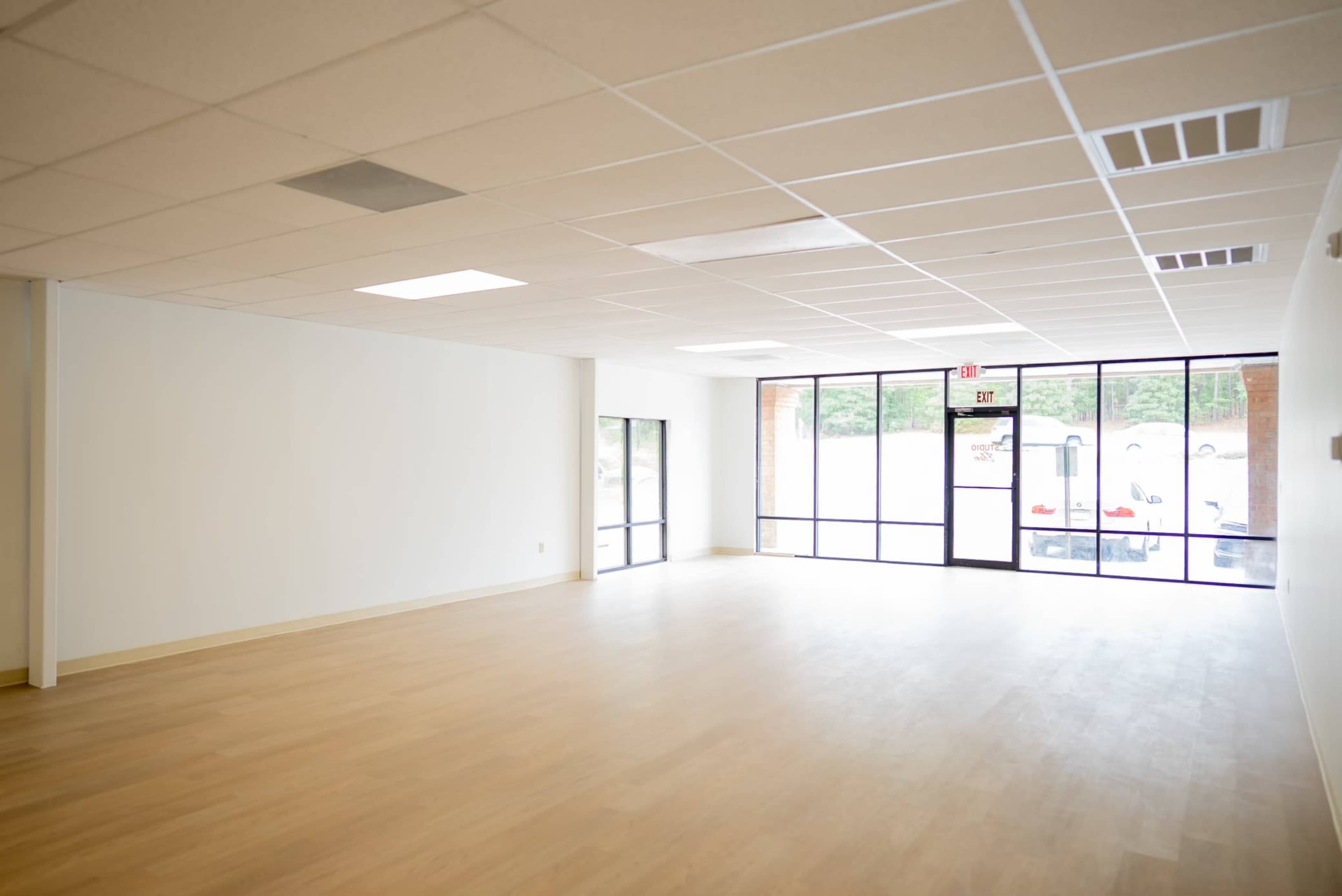 The image shows an empty commercial space with large windows, a light-colored floor, and a ceiling with acoustic tiles.