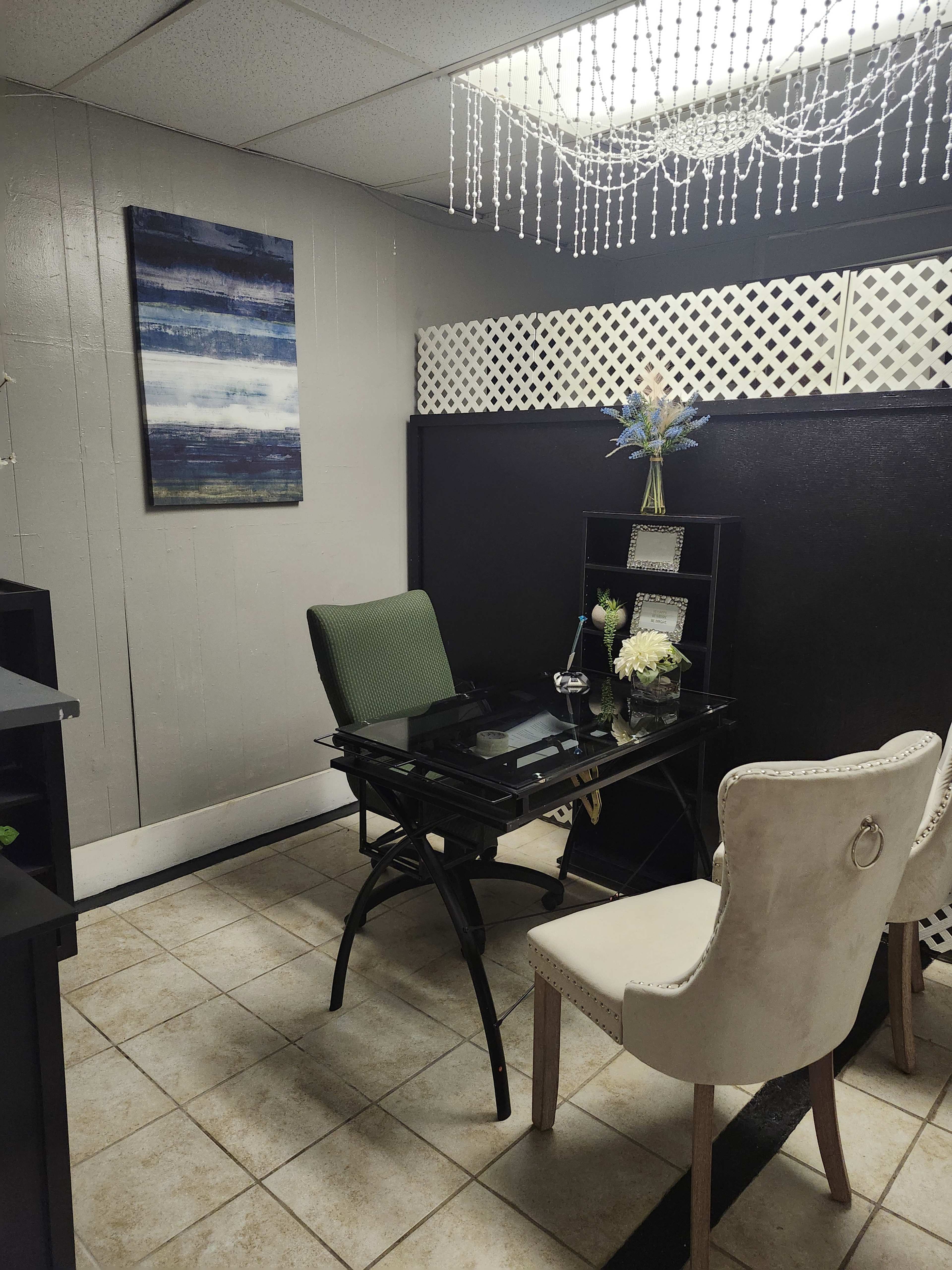 The image shows a small office space featuring a glass desk with a green chair and two white upholstered chairs, decorated with a piece of abstract art and a light fixture made of beads.