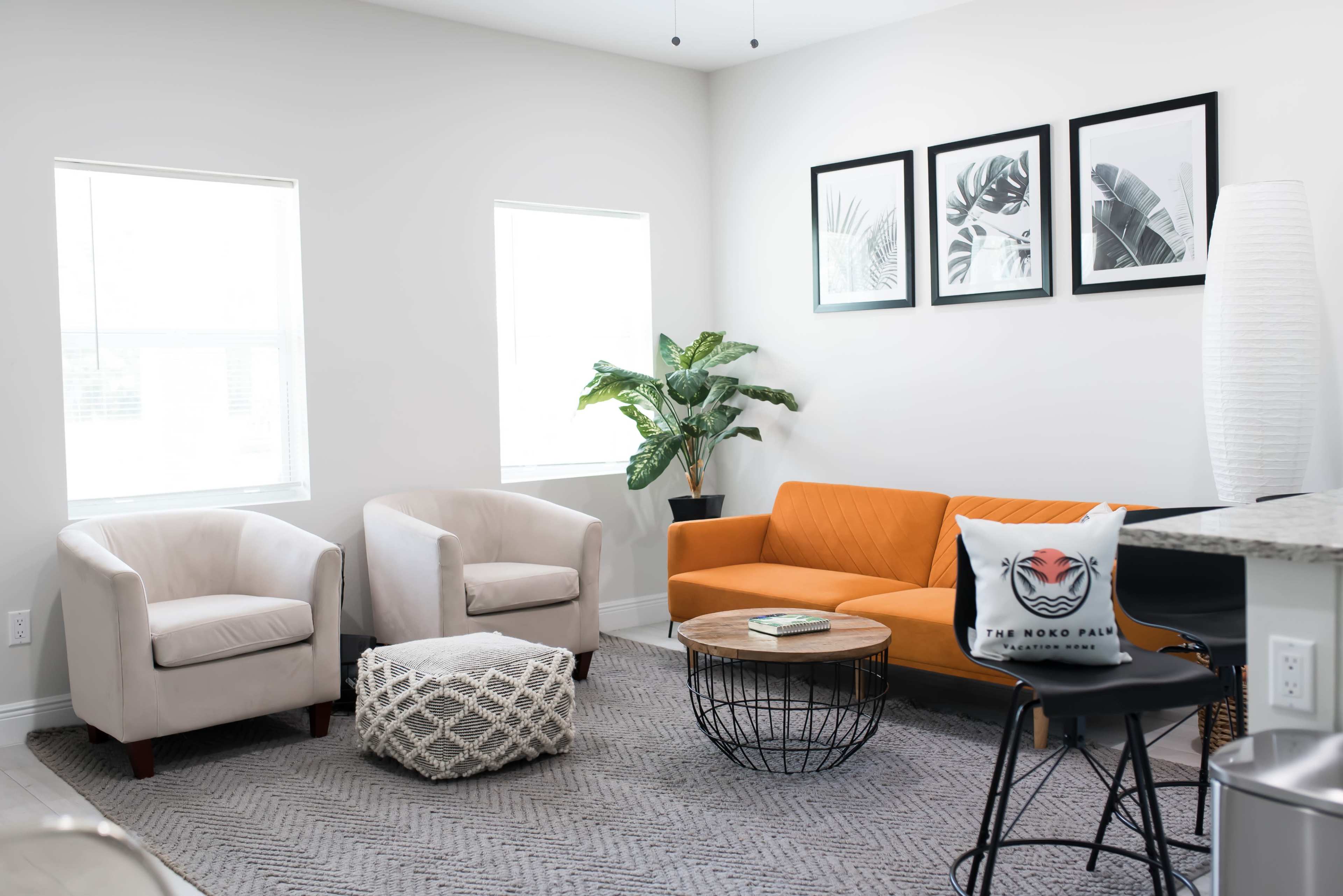 A modern living room with an orange sofa, two light-colored chairs, a wooden coffee table, a patterned rug, and potted plants, illuminated by natural light through two windows.