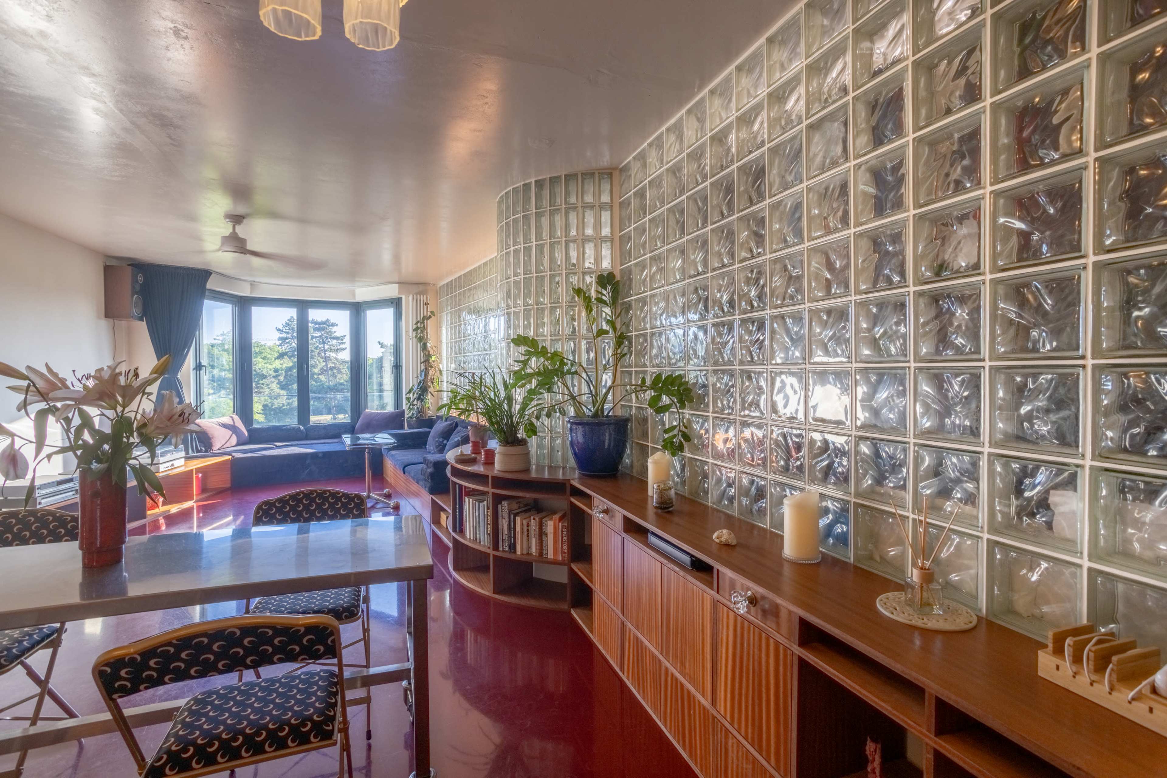 The image shows a modern interior space featuring a glass block wall, a wooden sideboard, and plants, with a view of the outdoors through large windows.