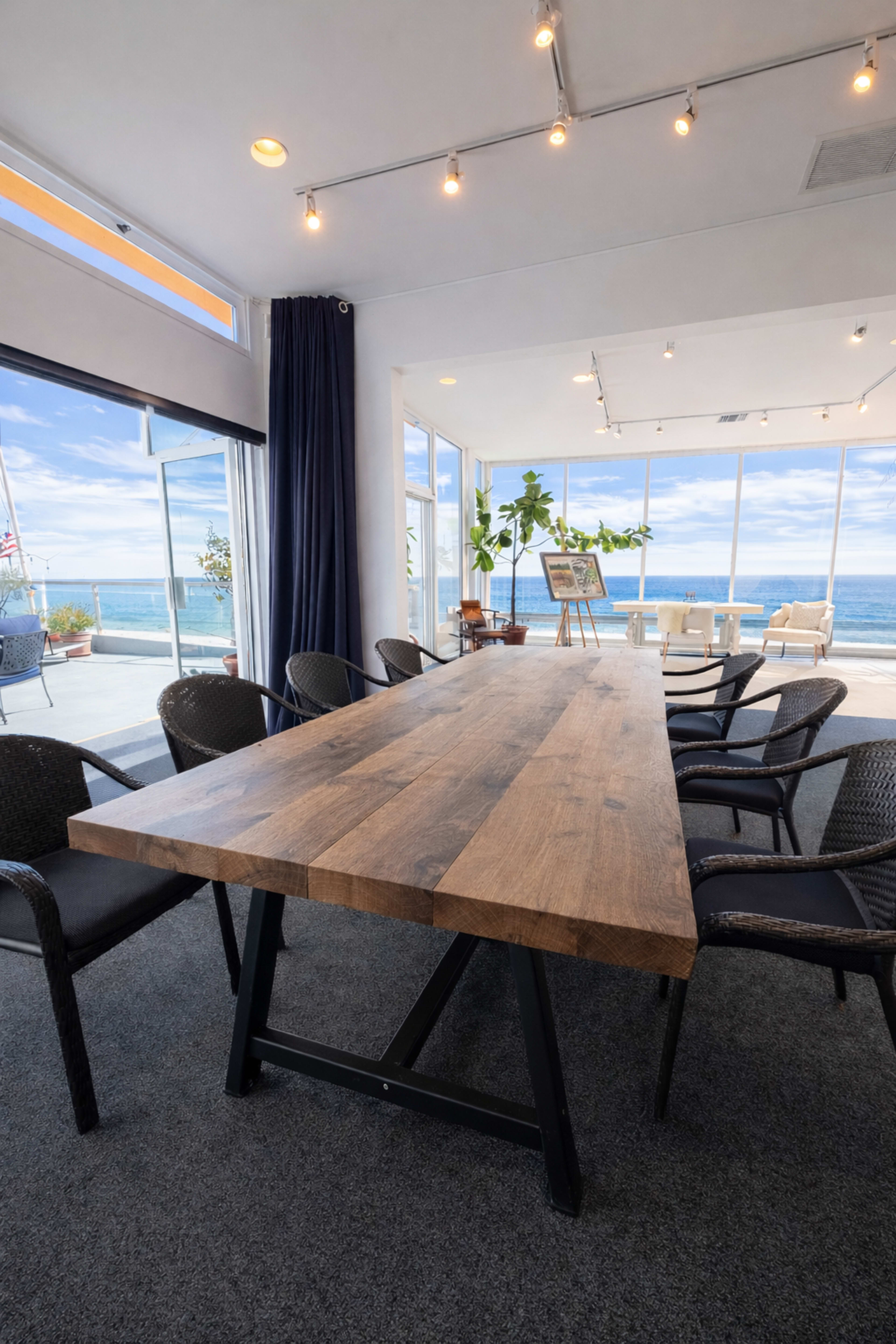 A long wooden table surrounded by chairs is positioned in a bright room with large windows overlooking a scenic view of the ocean.