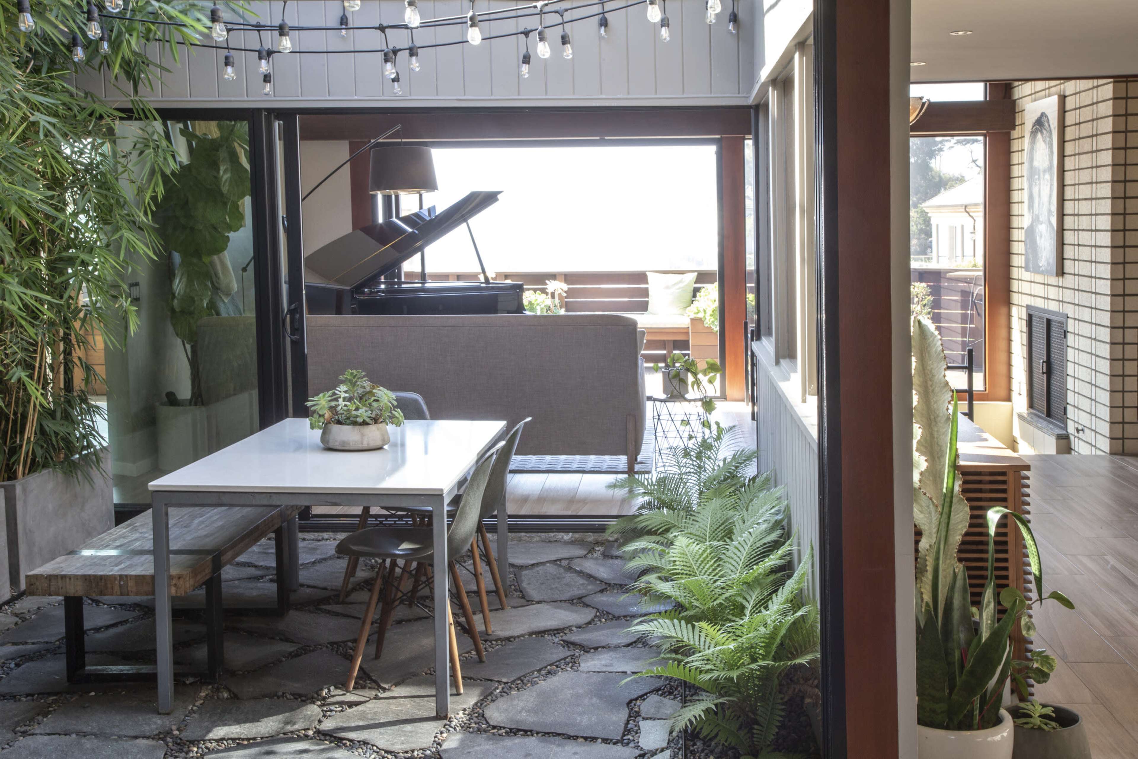 The image shows a modern indoor space featuring a dining table and chairs, surrounded by plants and stone path, with a piano visible in the background.