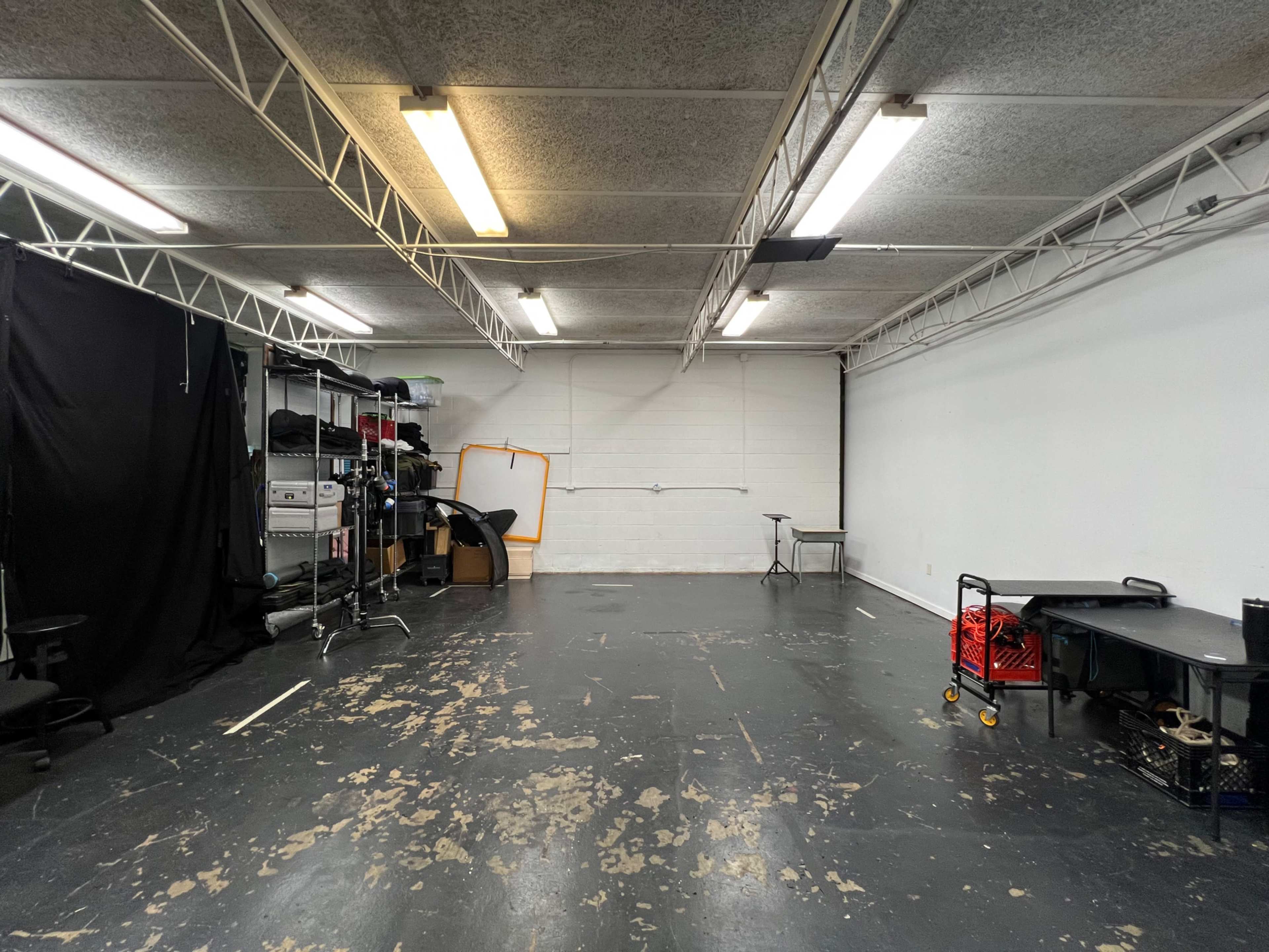 The image shows an empty studio space with overhead lighting, black curtains on one side, and various pieces of equipment and furniture along the walls.