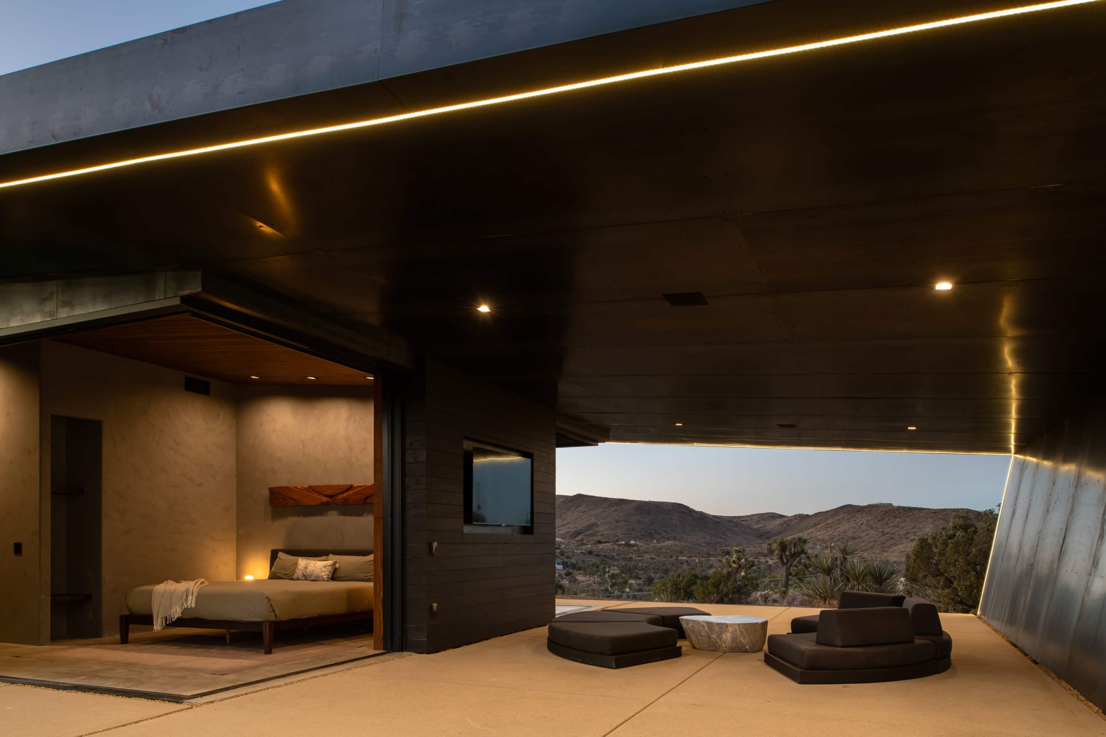 A modern, open-air living space features a bed and seating area, with a view of rolling desert hills under evening lights.