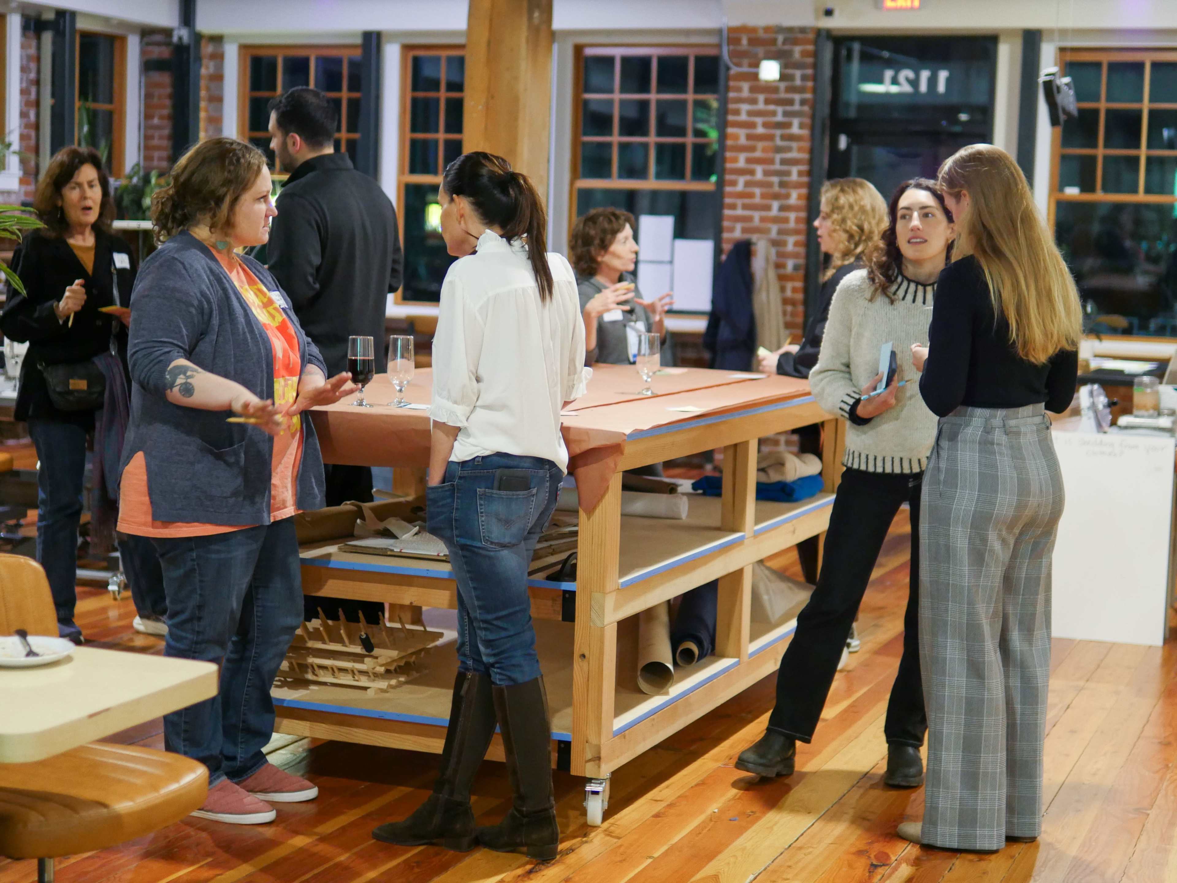 A group of people socializes around a large table in a well-lit room with wooden floors and brick walls.